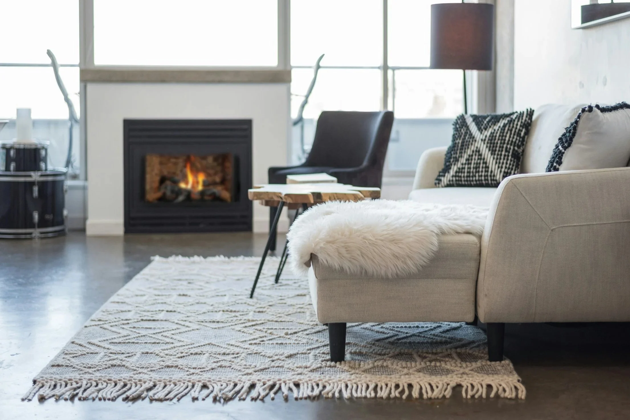 Living room with fireplace, beige sofa with black and white pillows, white fluffy throw, wooden coffee table, black armchair, rug, and large windows.