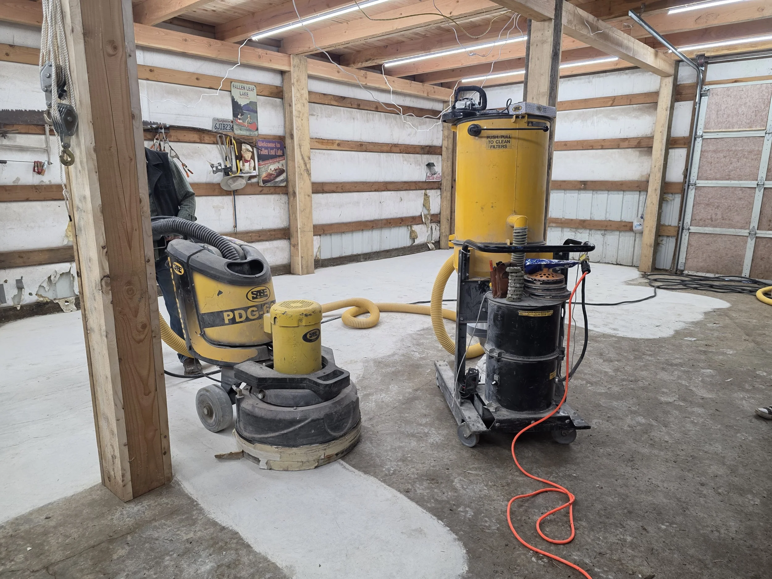 A garage or workshop space with unfinished walls and concrete floor. Two industrial sanding or polishing machines are present, connected with hoses and cords. Wooden framing and wall insulation are visible, along with various tools hanging on the wall.