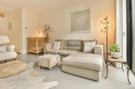 Living room with a beige sectional sofa with pillows, a small ottoman, floor lamp, potted plant, and a framed picture on the wall.