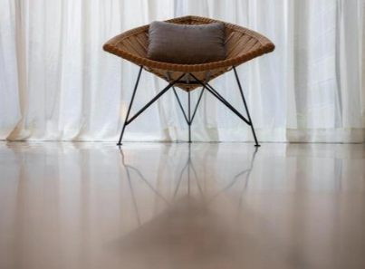 A modern chair with a round woven rattan back and seat, a dark cushion, and black metal legs, positioned in front of sheer white curtains.