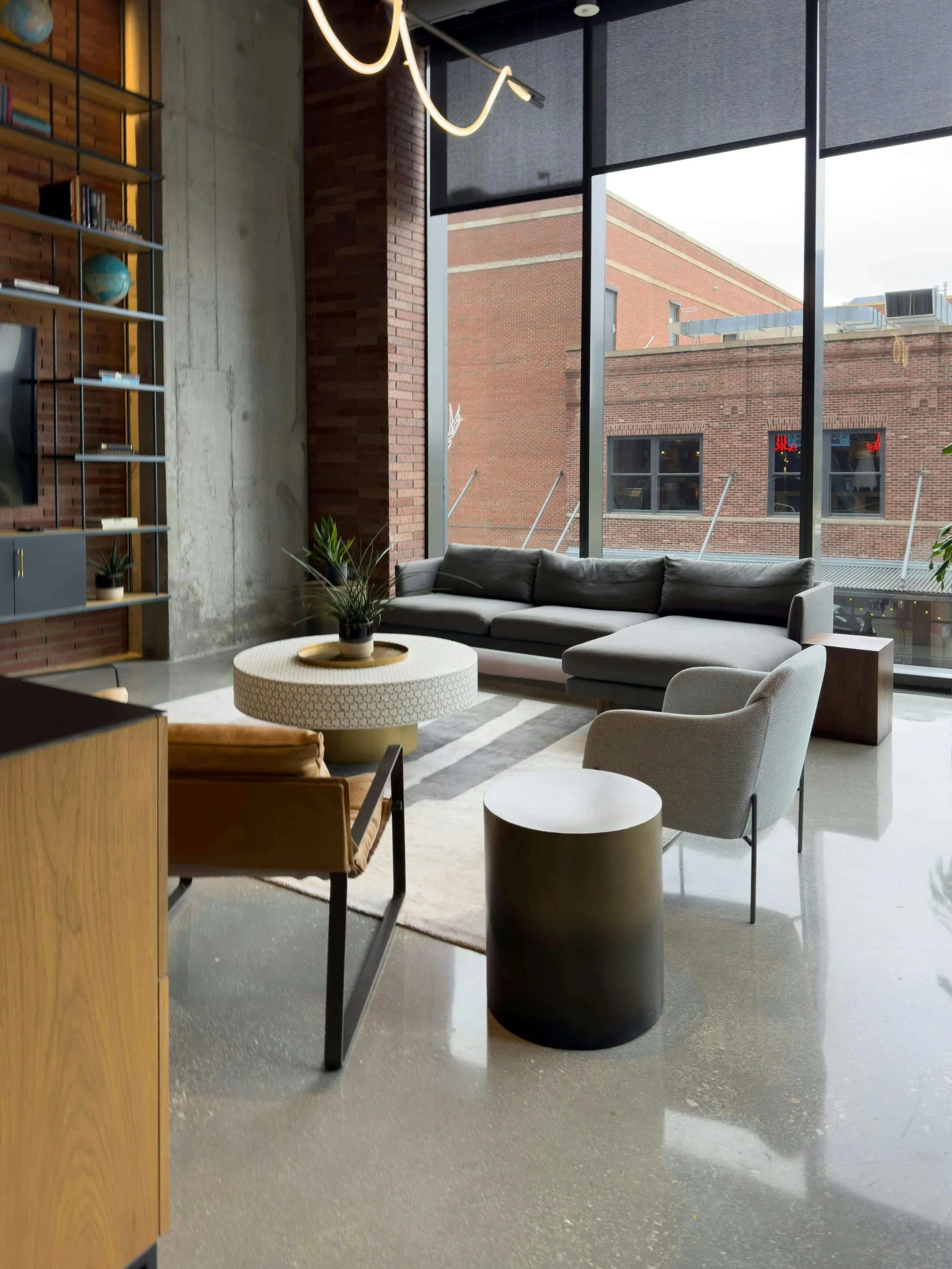 Modern urban living room with large floor-to-ceiling windows, furnished with a gray sectional sofa, a light gray armchair, a round coffee table with a potted plant, and a bookshelf with books and decorative items against a brick and concrete wall.