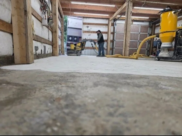 Person working in a garage or workshop with exposed wooden beams, using equipment connected to a yellow hose, with a concrete floor and tools on the wall.