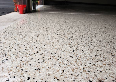 Close-up of a speckled concrete floor in a garage or warehouse, with some buckets and equipment in the background.