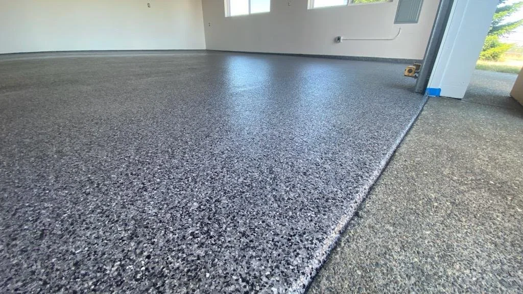 Smooth polished gray and black speckled epoxy garage floor with a clear edge transition to a rough gray concrete surface near the garage door.