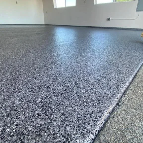 Empty room with speckled gray flooring and white walls, two windows letting in natural light, and an electrical outlet near the floor.