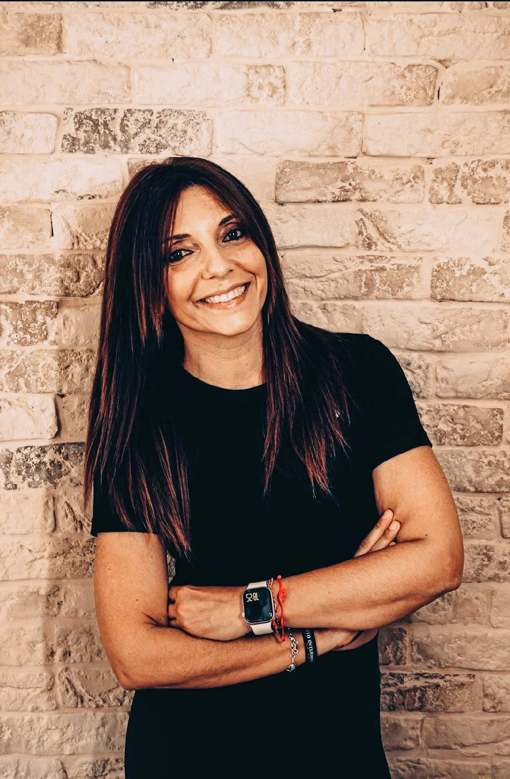 Mujer con cabello largo y oscuro, sonriendo, de pie frente a una pared de ladrillos beige, con brazos cruzados, vistiendo una camiseta negra, usando reloj inteligente y pulseras.