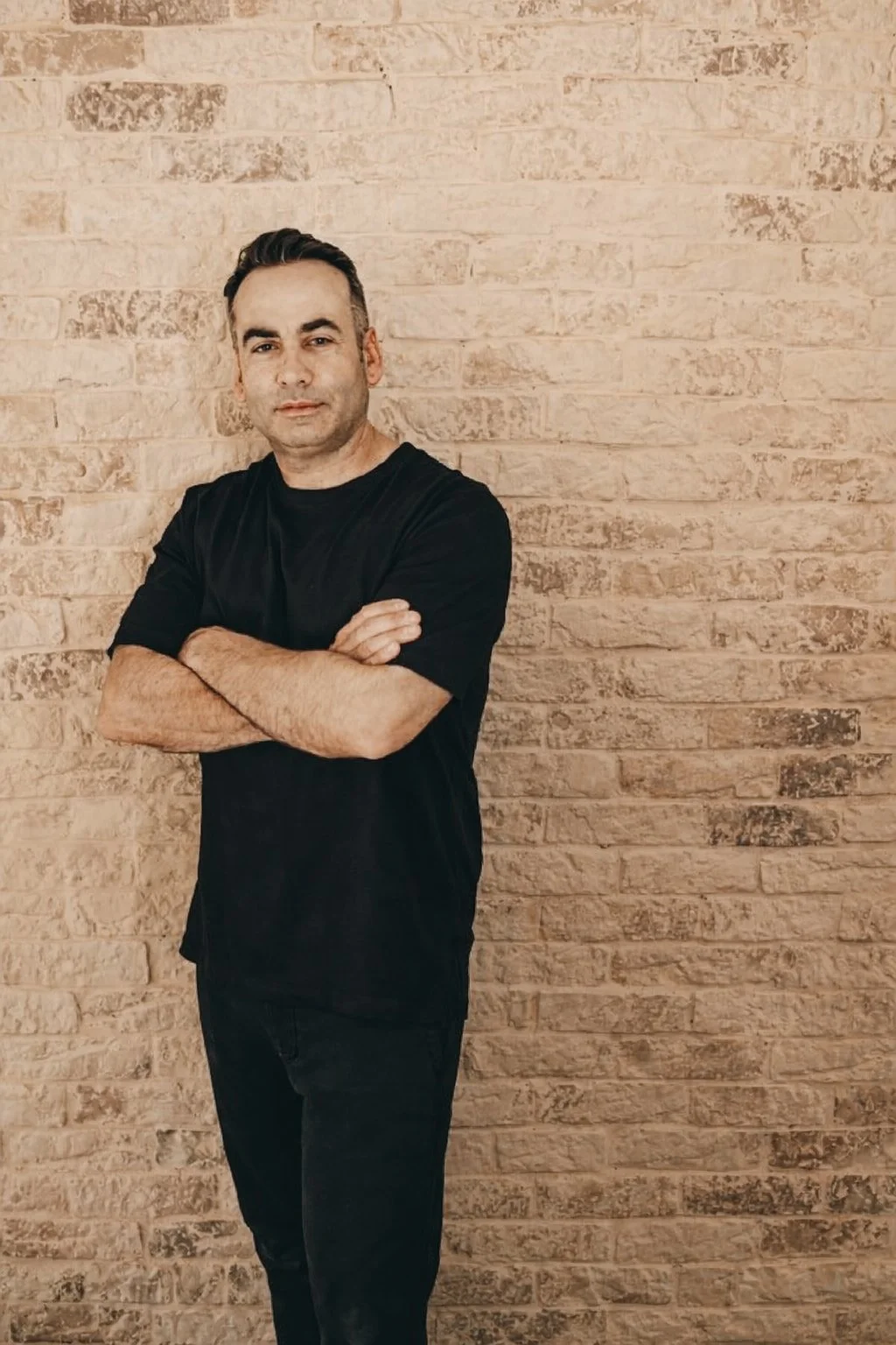 Un hombre de cabello oscuro y cejas gruesas, con piel clara, vestido con camiseta negra y pantalones oscuros, cruza los brazos y mira hacia la cámara, parado frente a una pared de ladrillos beige.