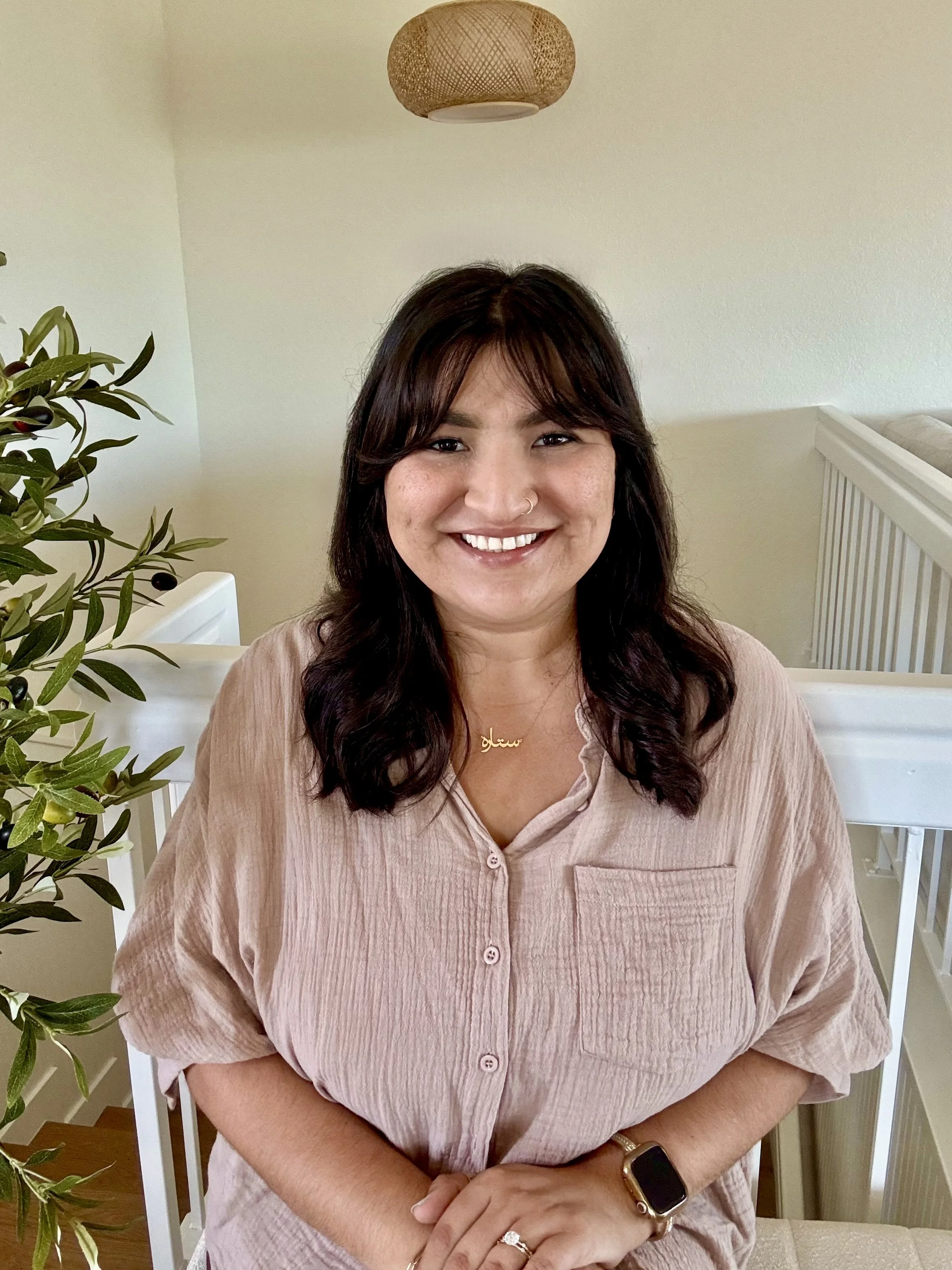 A woman with dark brown hair and light skin smiling, wearing a beige button-up shirt, a gold necklace, and a smartwatch, sitting in a bright room with a plant and a white railing in the background.