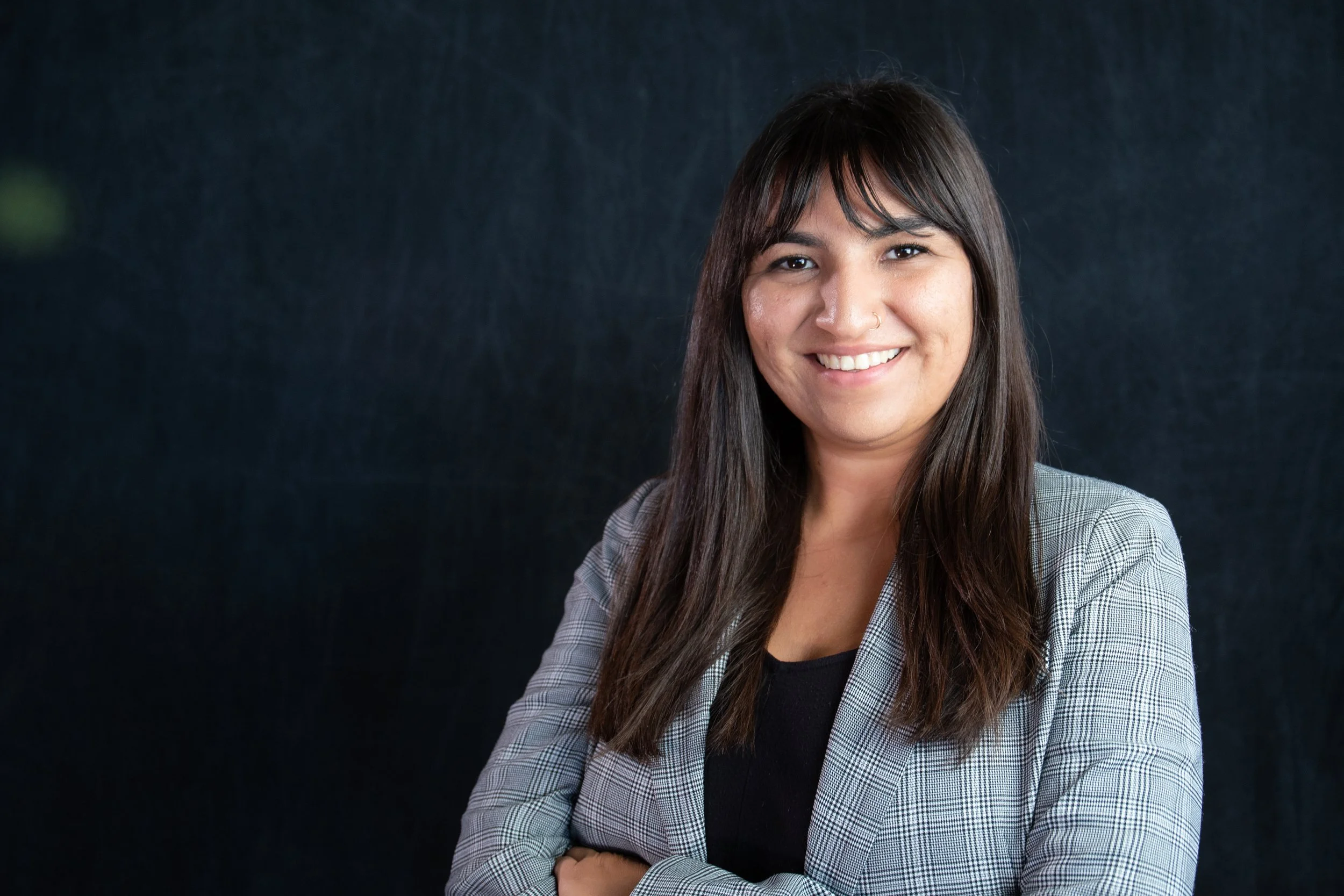 A woman with medium skin tone and long dark brown hair smiling with crossed arms, wearing a light gray blazer and black shirt, standing against a dark background.