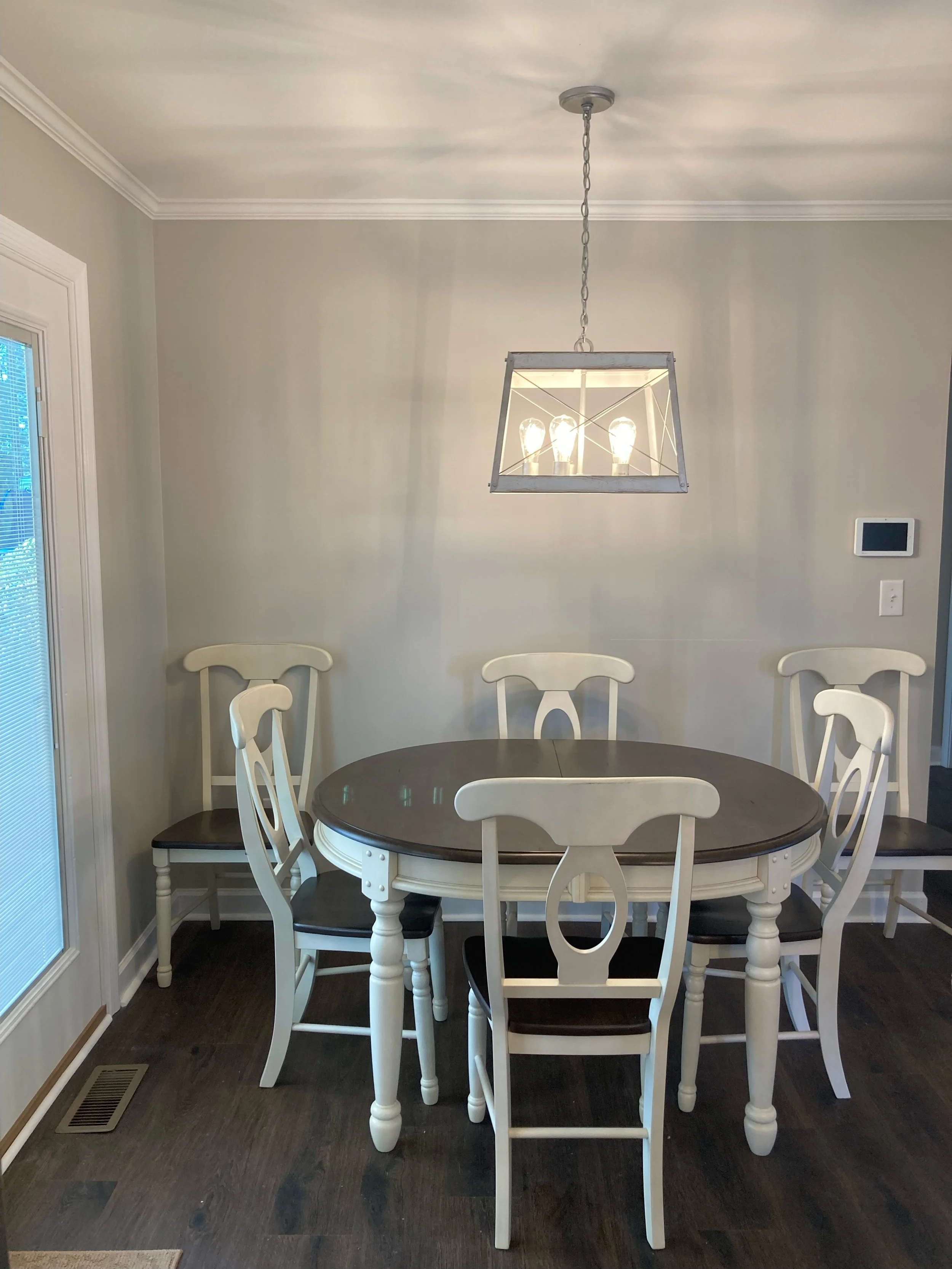 White dining table surrounded by white chairs in a room with beige walls and a hanging light fixture with three light bulbs.