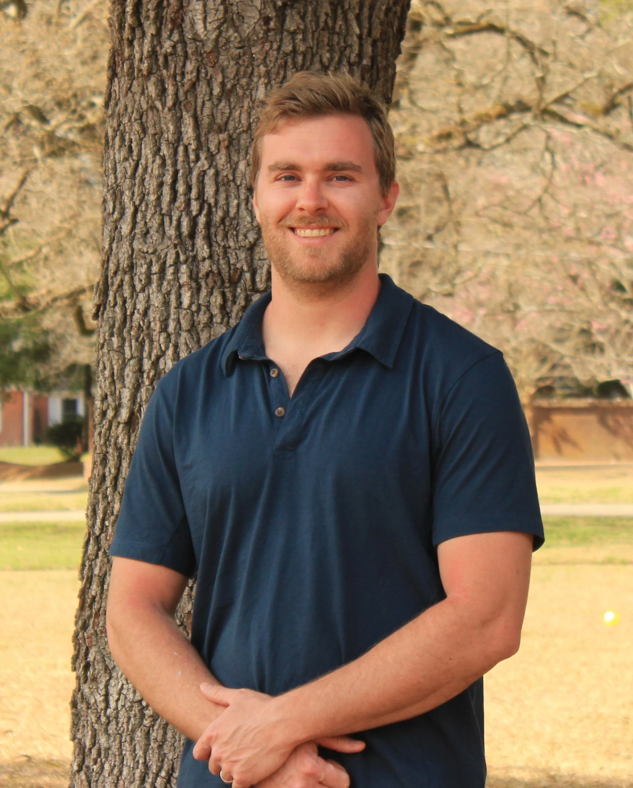 A young man with light brown hair and a beard smiling, standing outdoors next to a tree with a background of trees and grass.