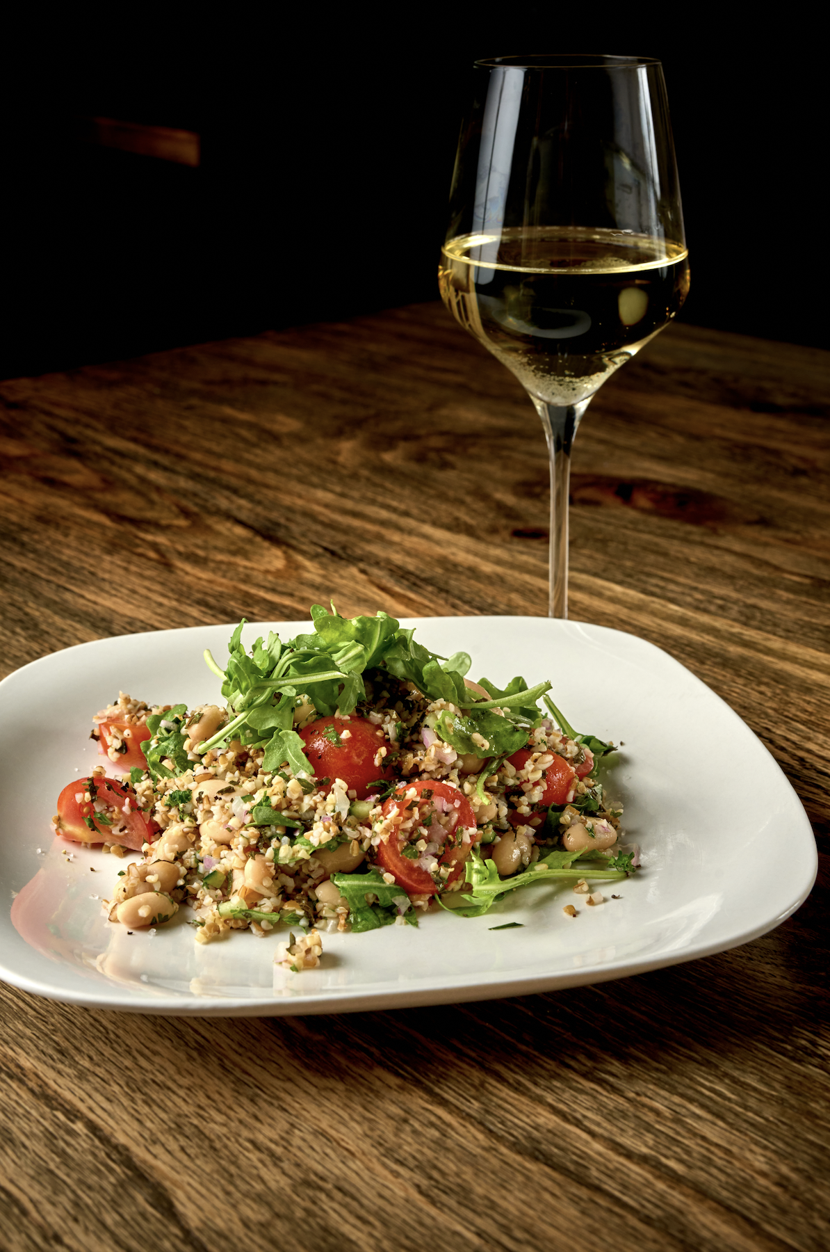 A plate of salad with cherry tomatoes, greens, and chopped nuts, next to a glass of white wine on a wooden table at night.