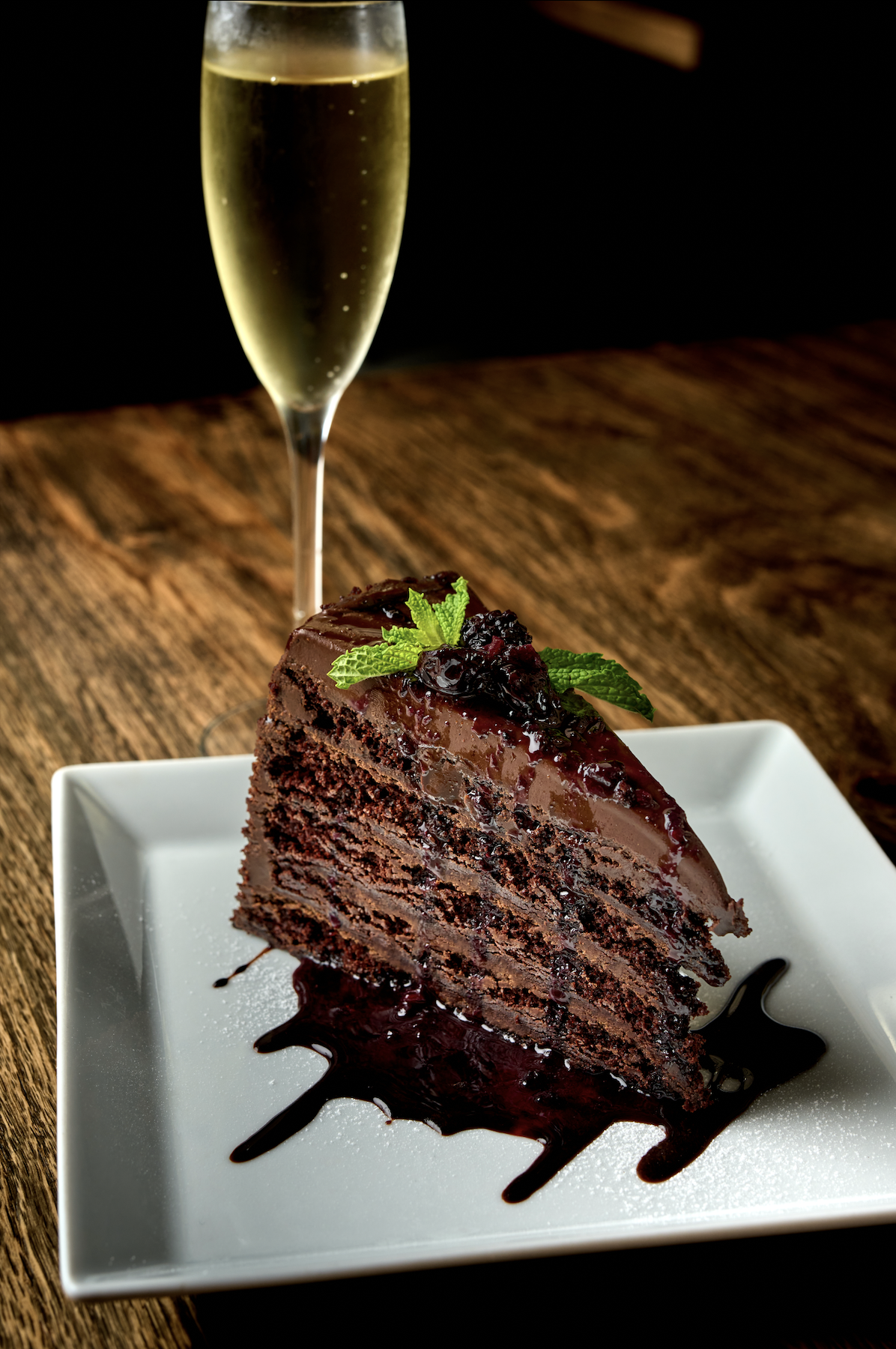 A slice of chocolate cake with berry topping and mint garnish on a white plate, with a glass of champagne in the background.