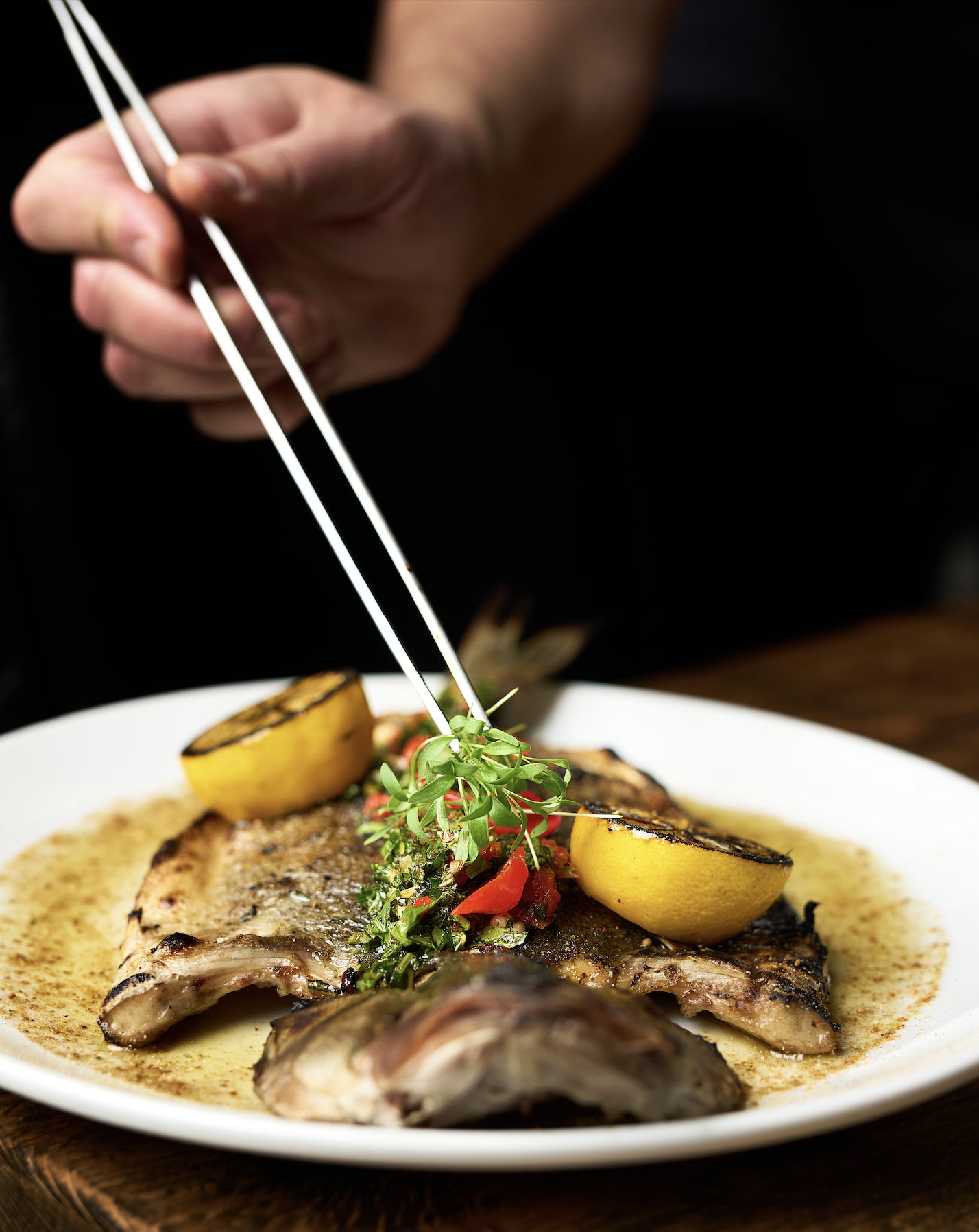 Close-up of a plated grilled fish dish garnished with herbs, lemon wedges, and a vegetable medley, with a person using tongs to add finishing touches.