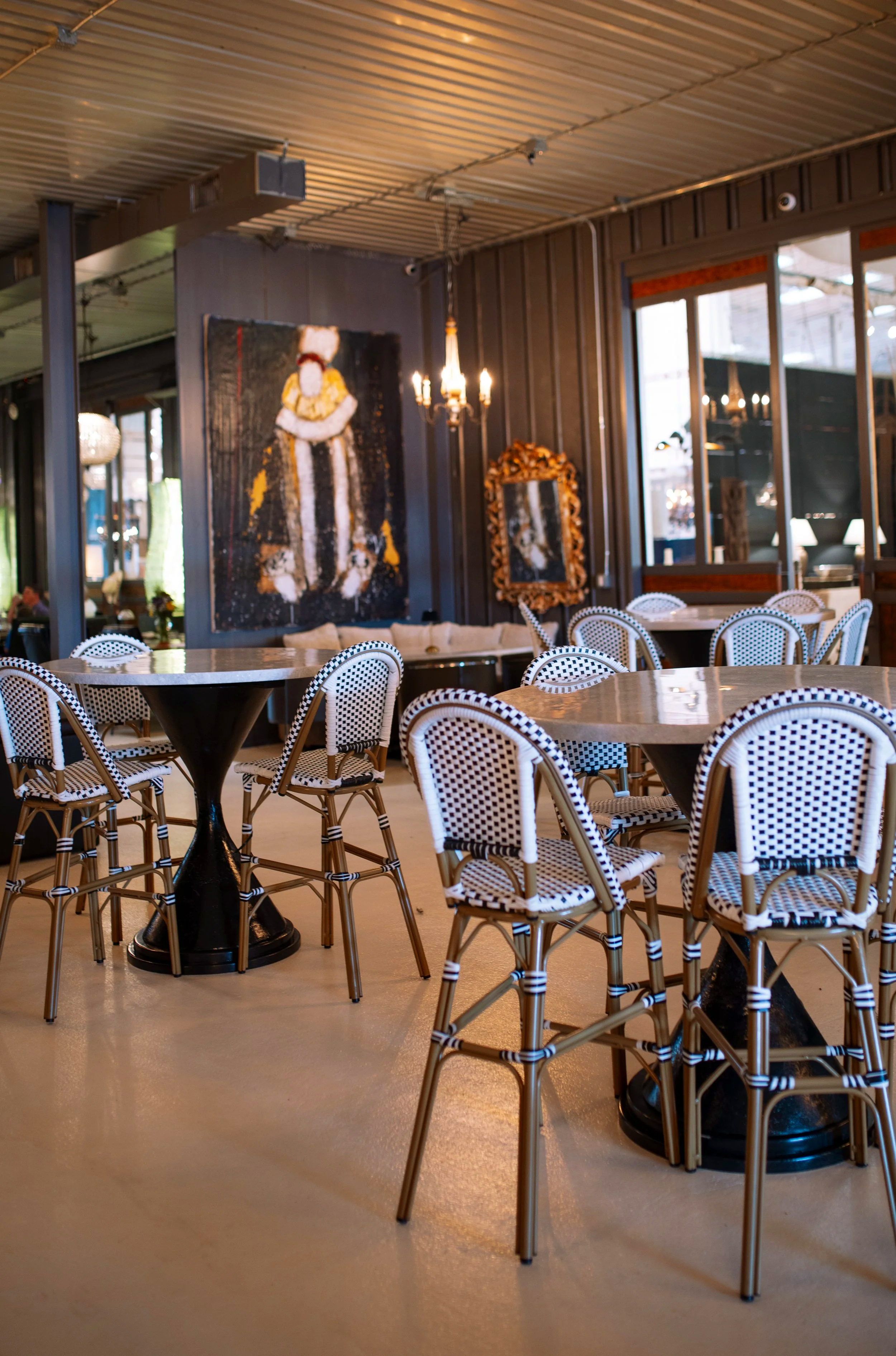 Interior of a restaurant or cafe featuring multiple round tables with black pedestal bases and black and white woven chairs. The wall behind has a large abstract painting and a decorative mirror with a gold ornate frame. Large windows let in natural light, and a chandelier hangs from the ceiling.