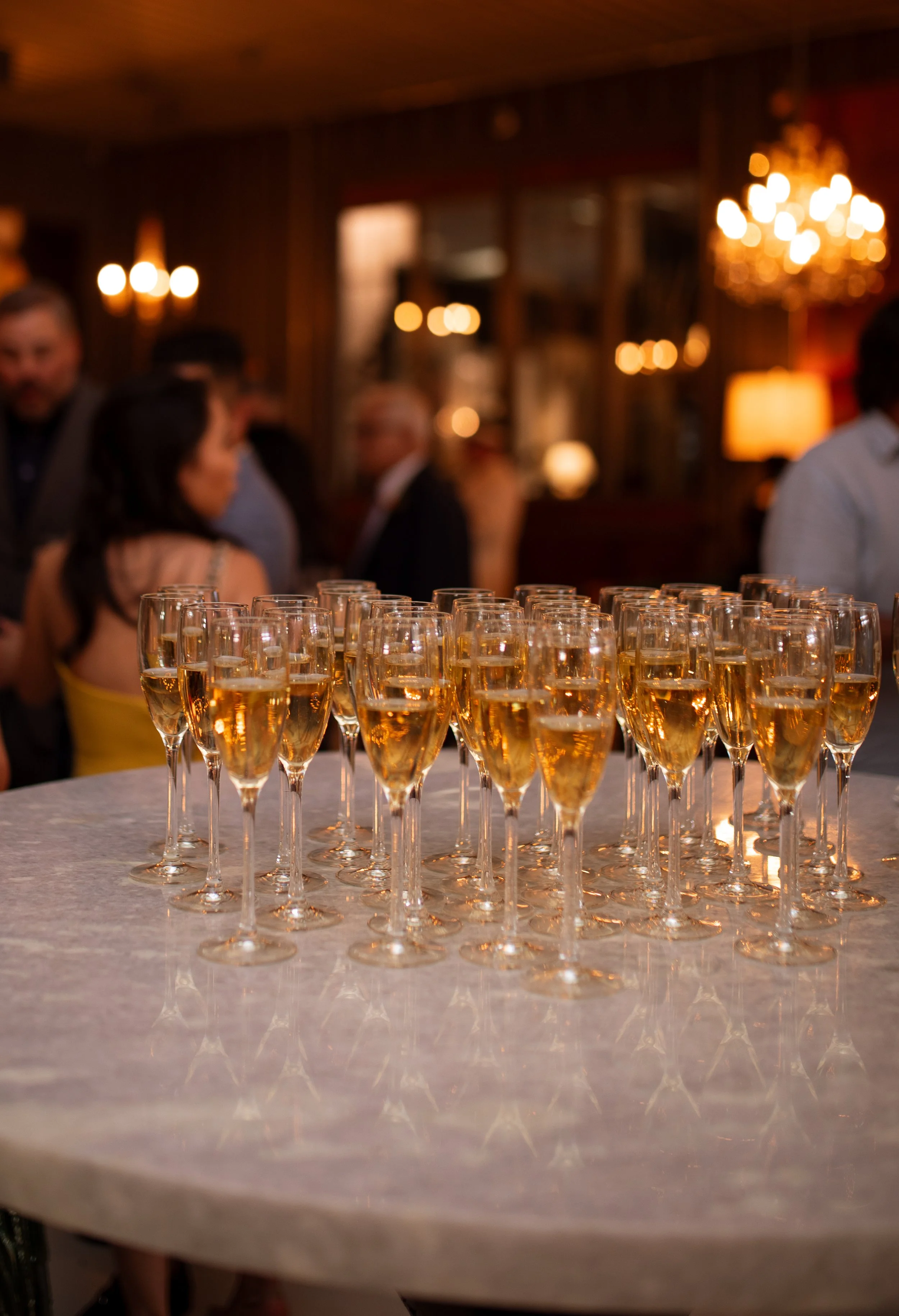 A table with multiple glasses of champagne in a dimly lit room with warm lighting and people in the background.