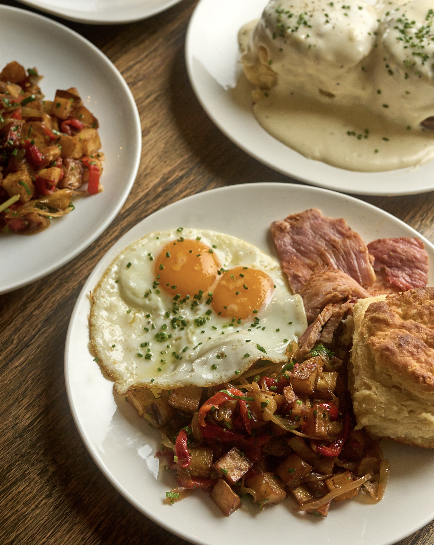 A breakfast plate with two fried eggs, bacon, sautéed vegetables, and a fried dish, possibly a potato pancake or bread