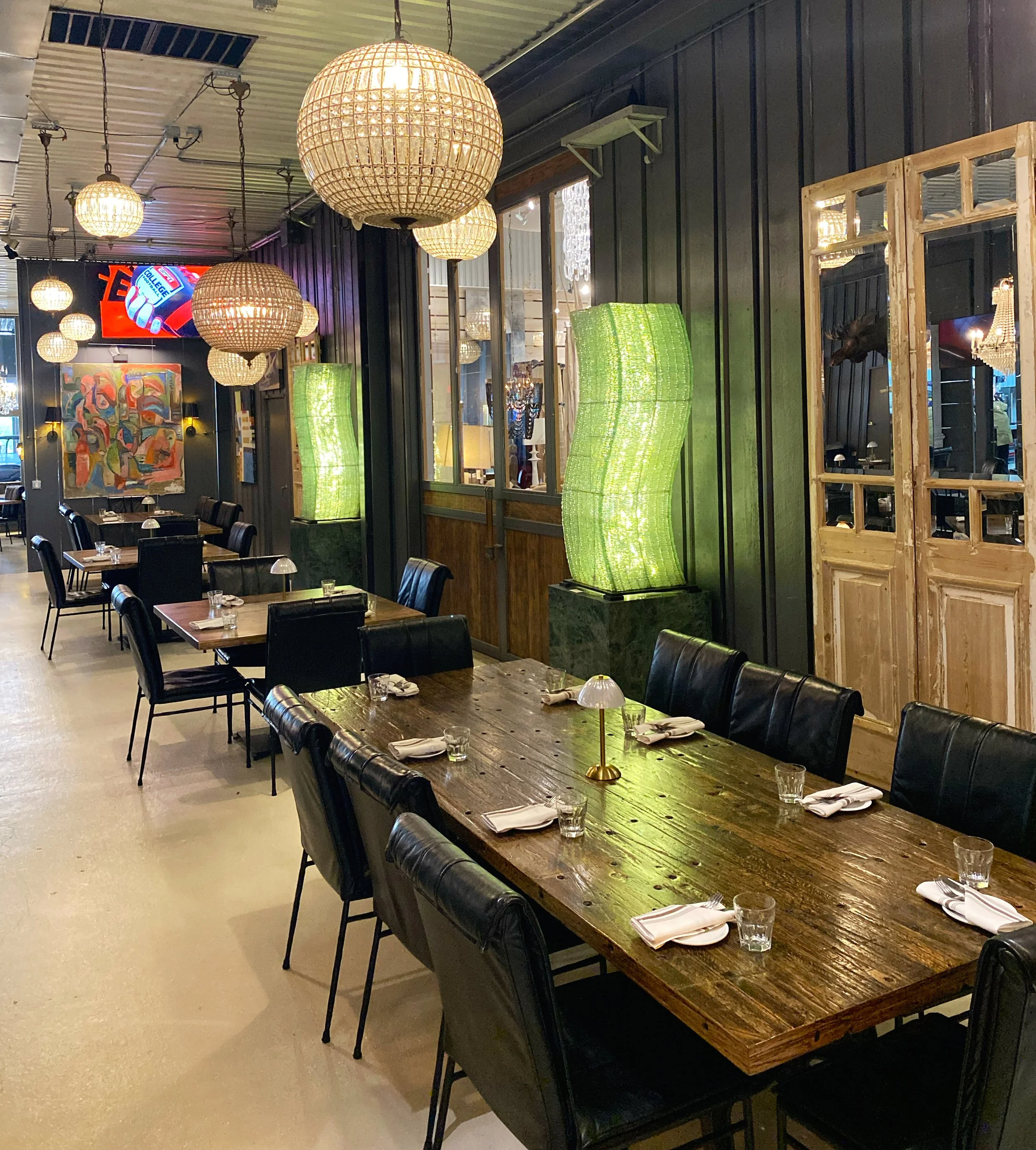 The image shows a dining area with a long wooden table set with napkins, glasses, and small lamps. There are multiple black chairs around the table. The background features large glass windows, green lamps, wooden frames, and decorative light fixtures hanging from the ceiling. Artwork and additional lighting are also visible.