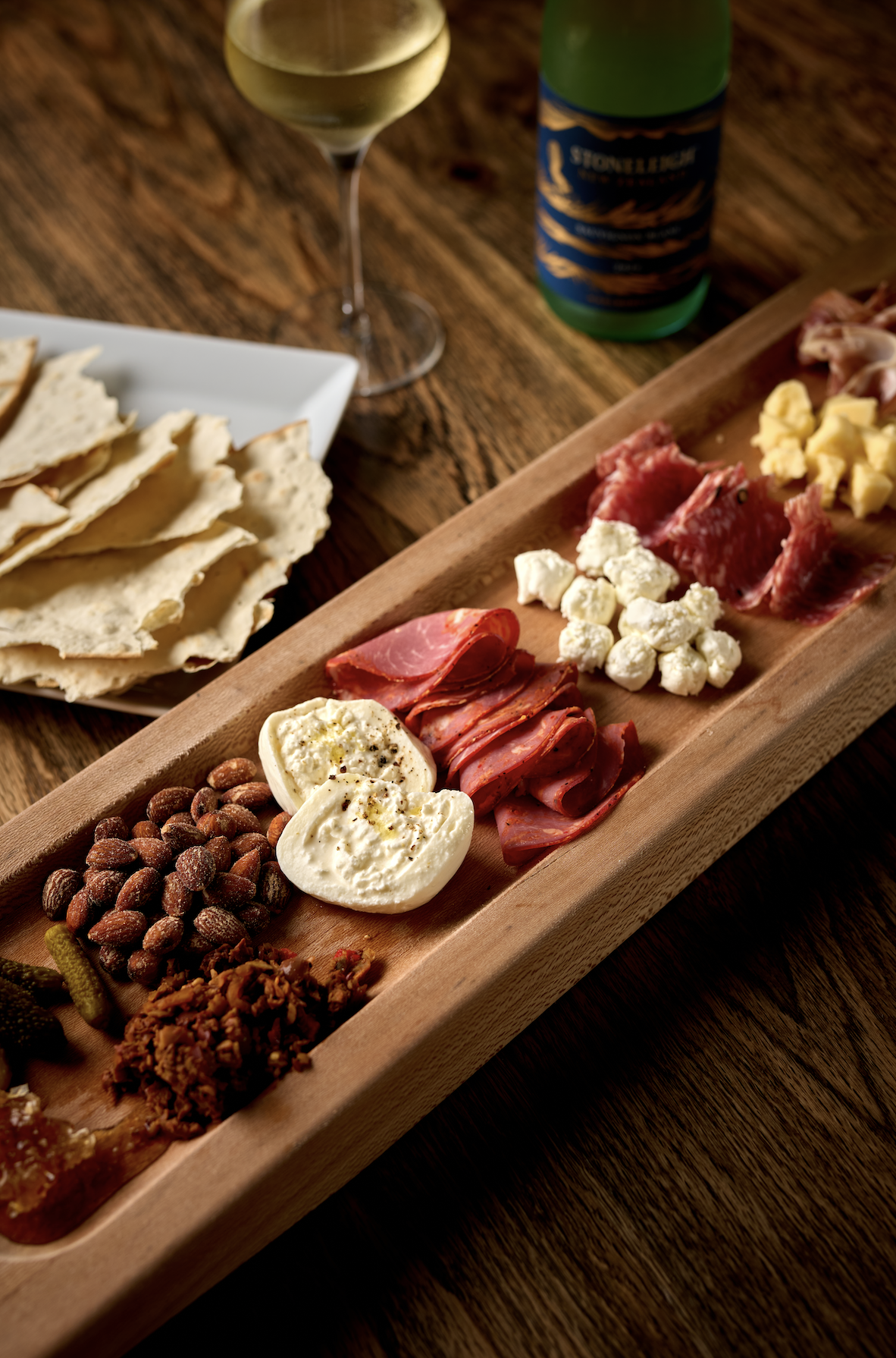 Charcuterie board with various cheeses, meats, nuts, and accompaniments, alongside a glass of white wine and a bottle of sparkling beverage on a wooden table.