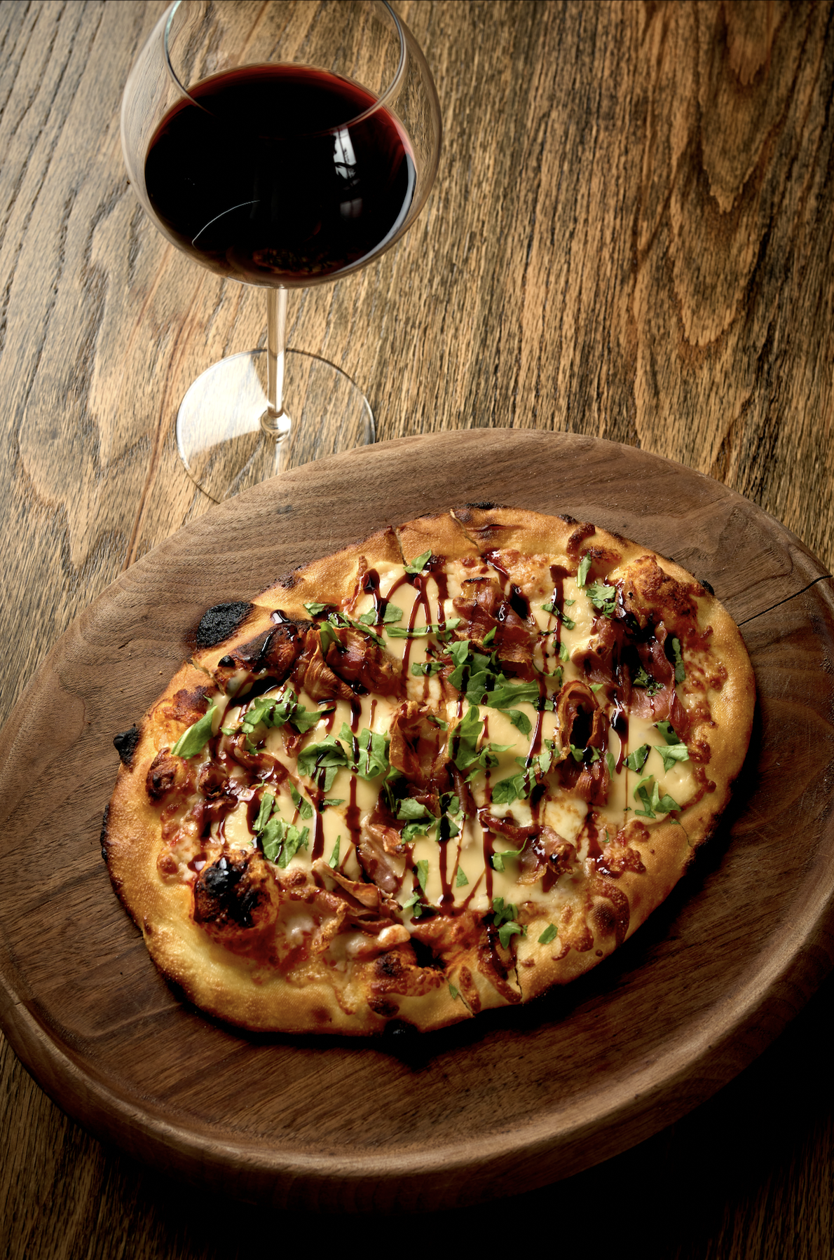 A glass of red wine next to a pizza topped with cheese, bacon, green herbs, and drizzled with balsamic glaze, on a wooden table.