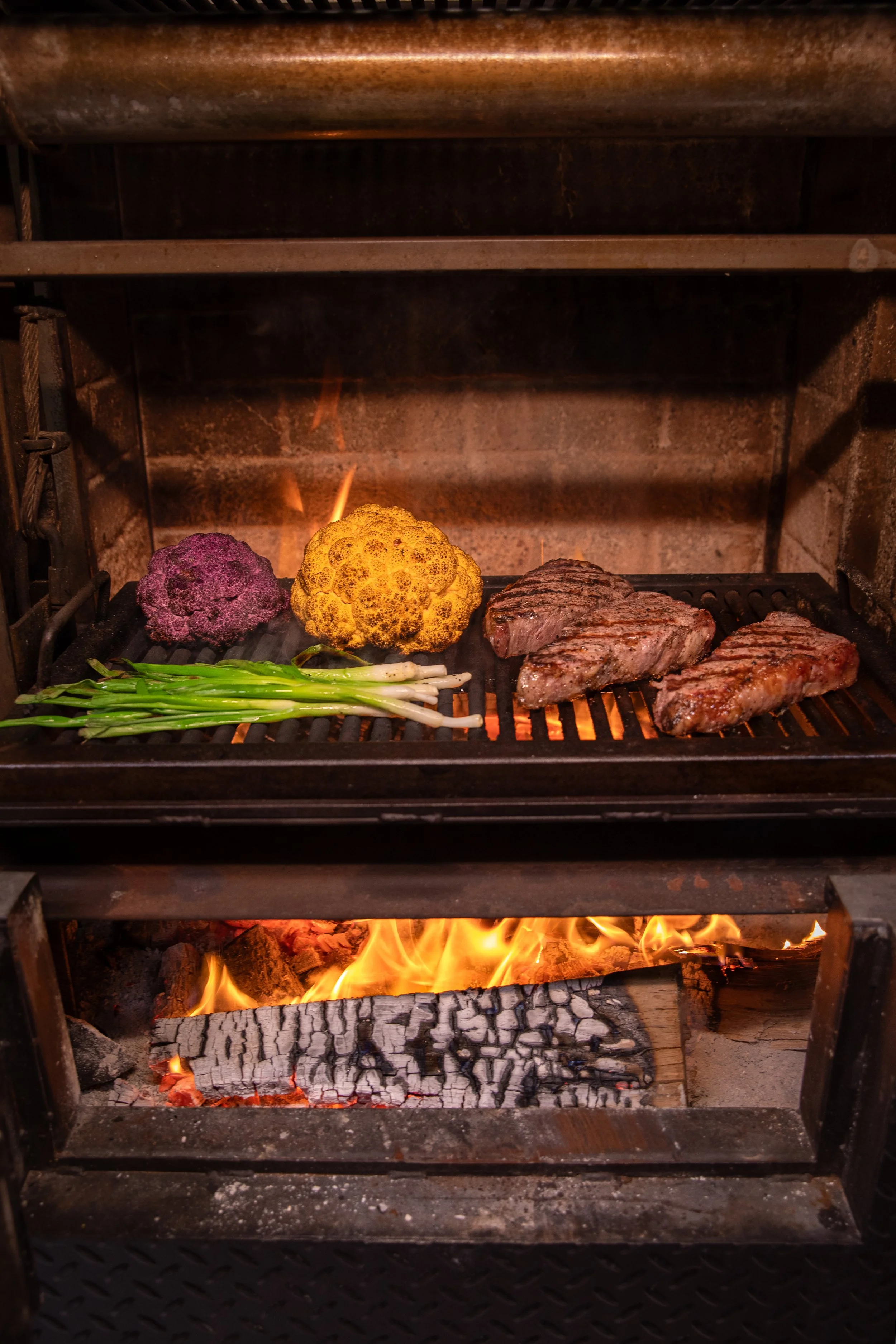 Beef steaks grilling with cauliflower, purple cauliflower, and green onions inside a barbecue grill with fire and charcoal