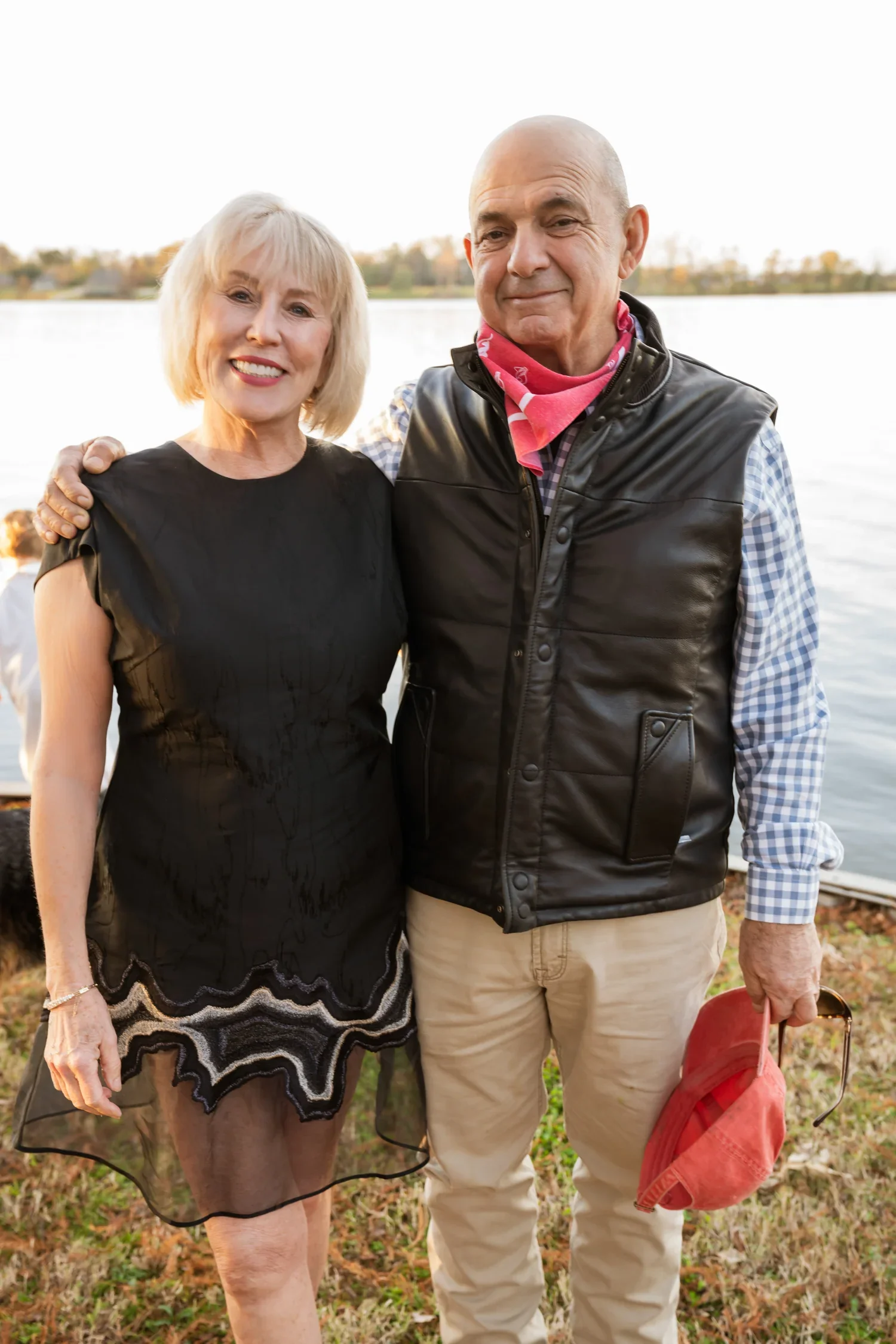 Older woman and man standing by a lake, smiling at the camera, with trees and water in the background during daytime.