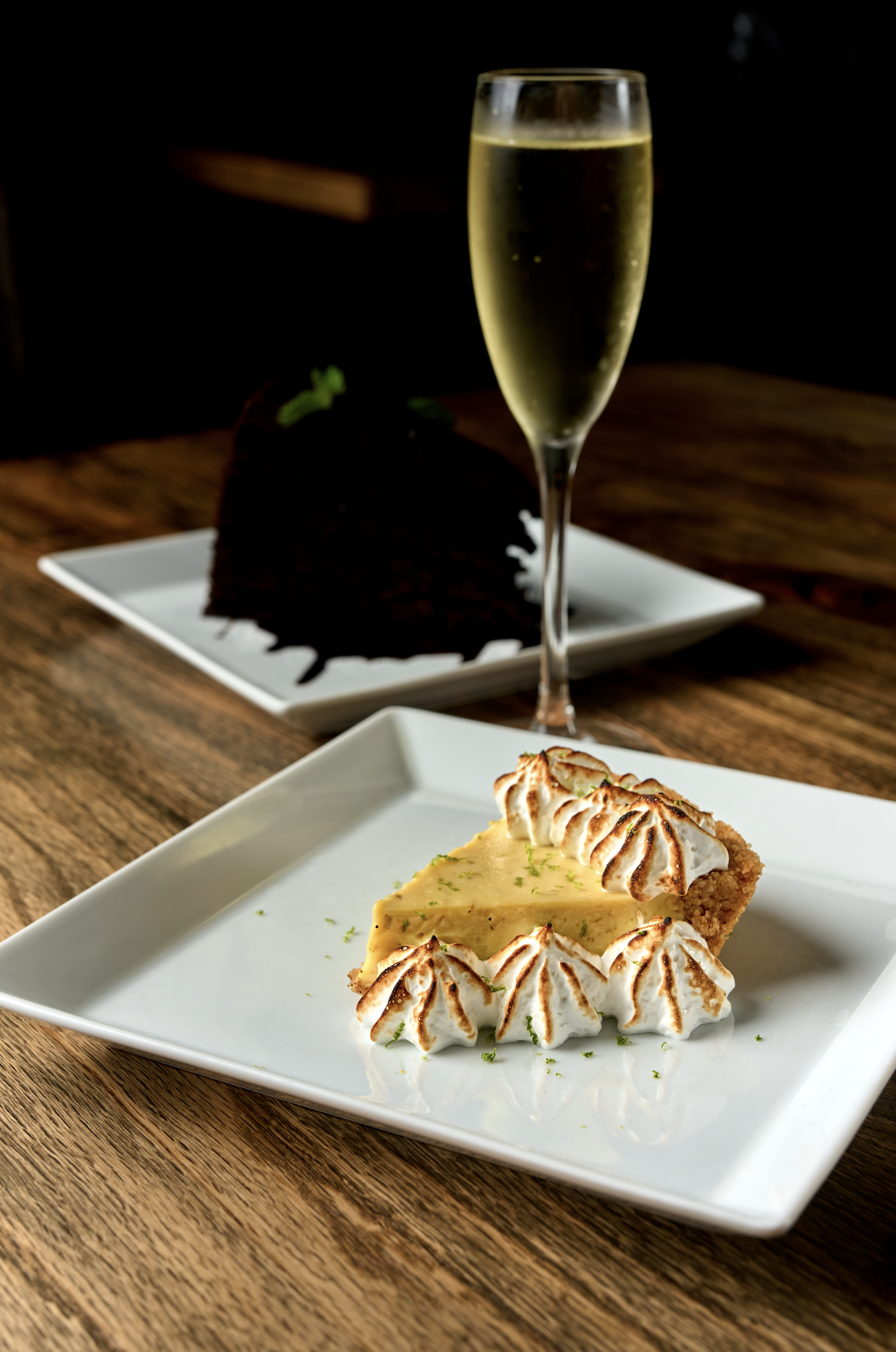 Slice of lemon meringue pie with toasted meringue peaks on a white plate in front, a glass of champagne behind, and a slice of chocolate cake on a separate plate in the background, on a wooden table.
