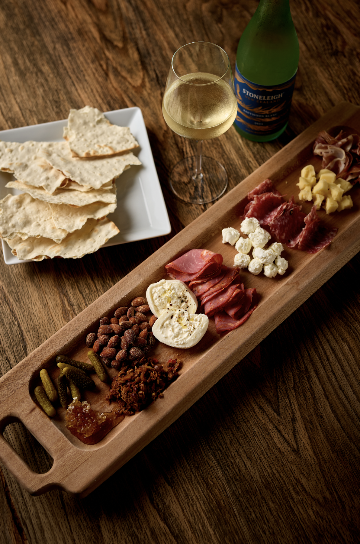 Charcuterie board with various cheeses, meats, nuts, and spreads, accompanied by a glass of white wine and a bottle of sparkling beverage, on a wooden table.