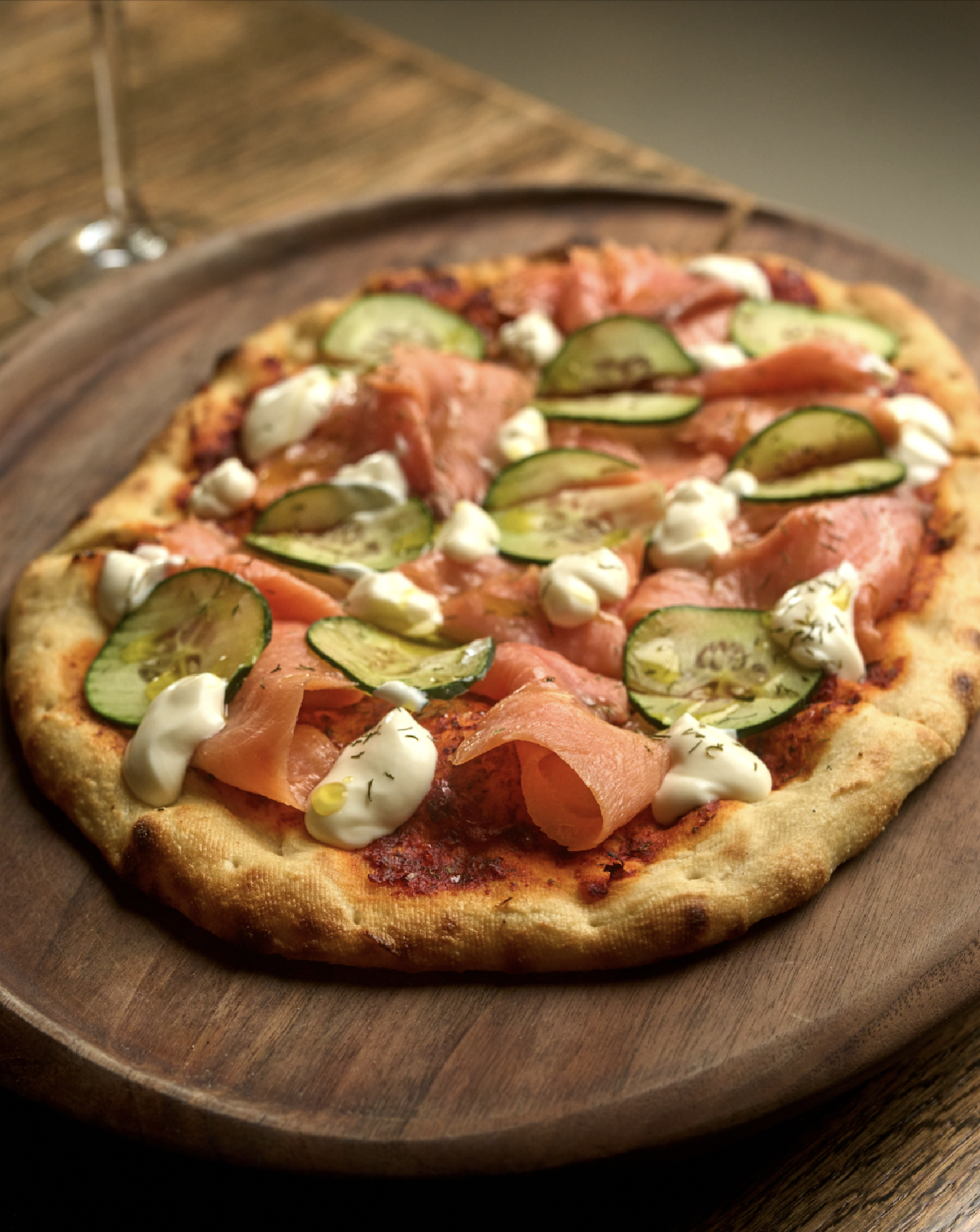A pizza with smoked salmon, cucumber slices, dollops of cream, and herbs on a wooden pizza board.