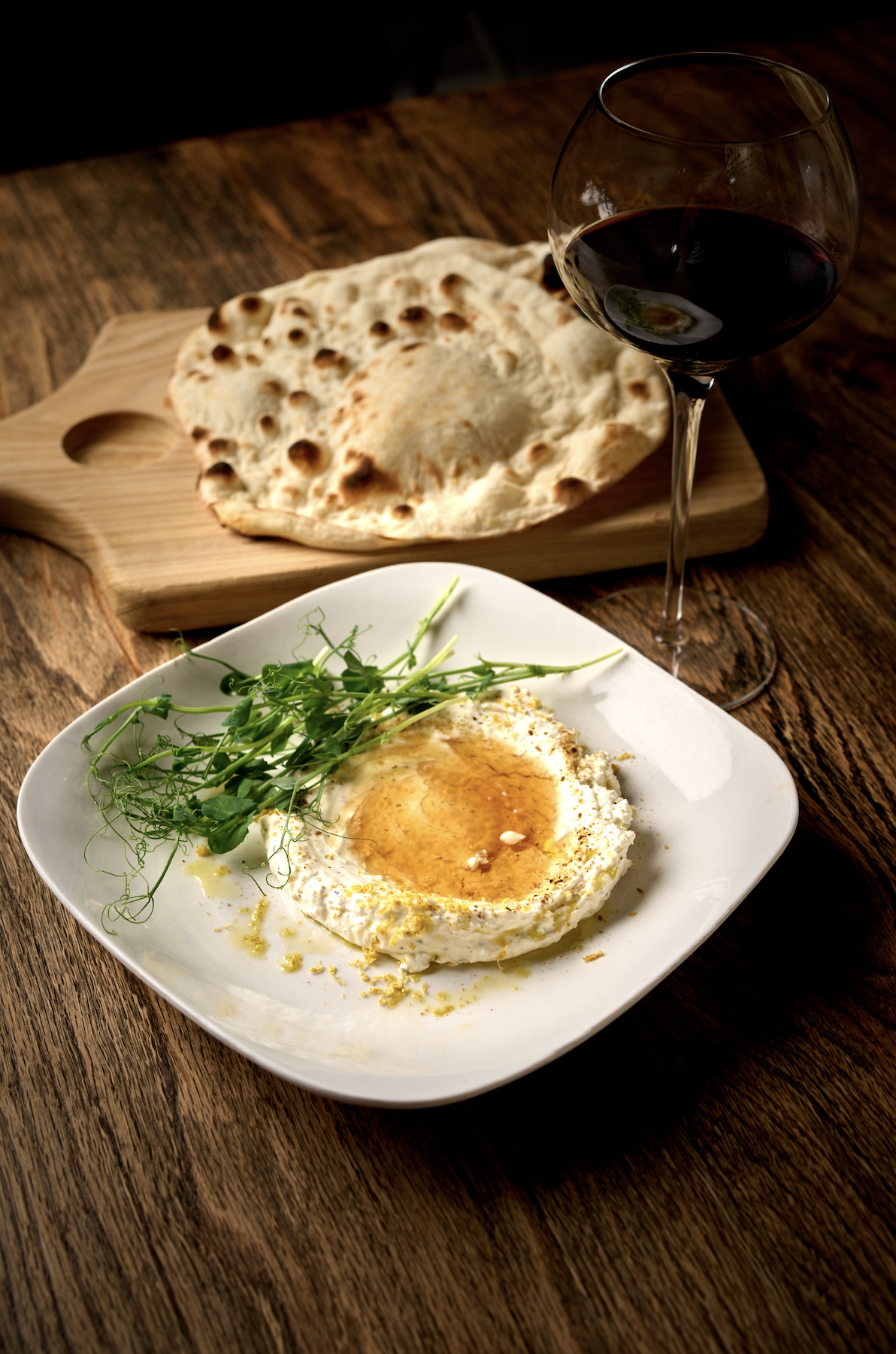 A plate with a fried egg topped with pepper and herbs, garnished with microgreens. A glass of red wine and a piece of naan bread on a wooden cutting board in the background.