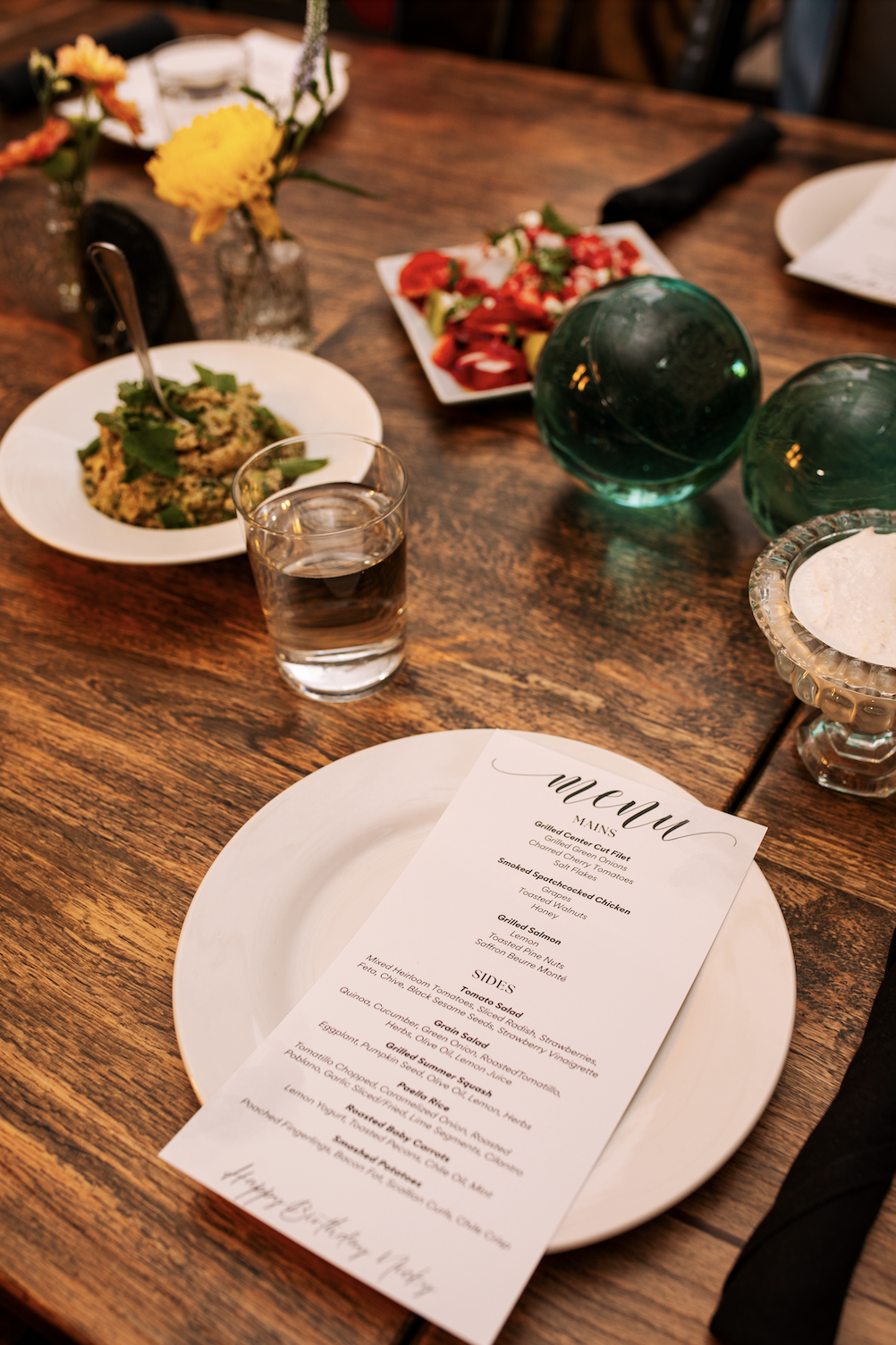 A wooden dining table set with a menu, a glass of water, dishes including guacamole, salad, and roasted vegetables, decorative glass globes, and flower vases.