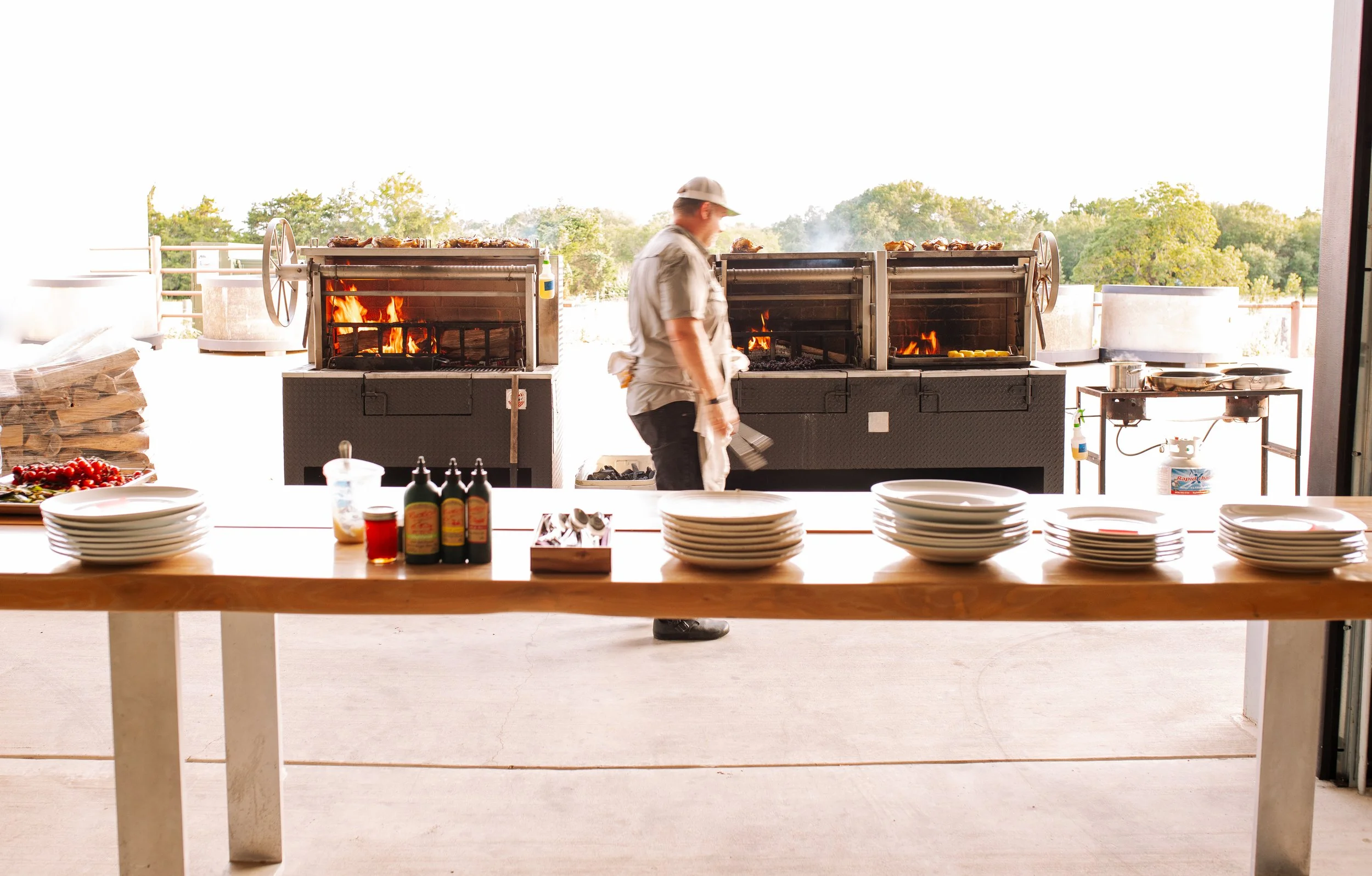 A man stands in front of outdoor grills cooking meat, with plates and condiments on a table in the foreground and trees in the background.