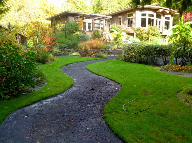 Luxury landscaping. A winding stone pathway leads through a well-manicured backyard with lush green grass, shrubs, and trees towards a house with large windows and a porch, surrounded by garden plants.