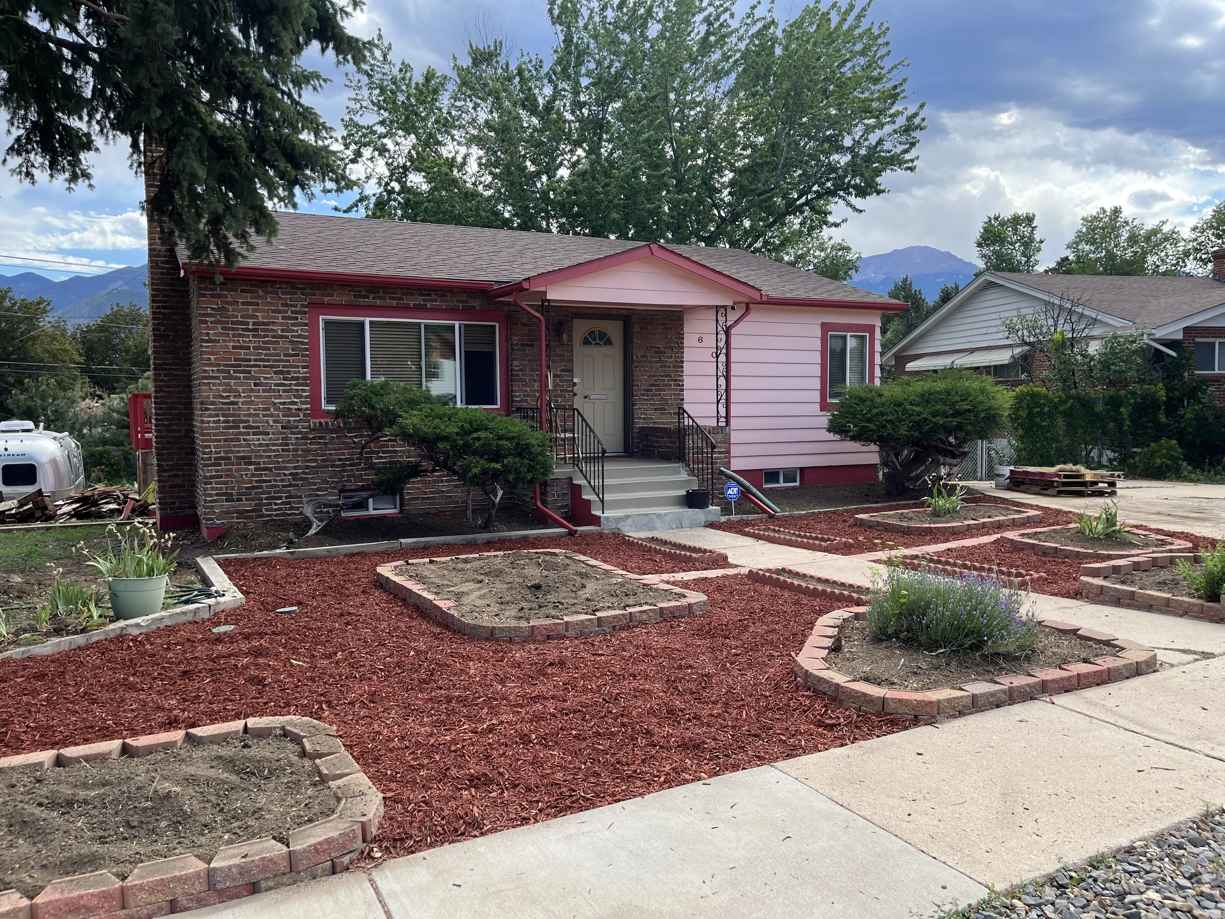 A house with a brick and pink exterior, landscaping with red mulch, small trees, and flower beds, along a concrete sidewalk, setting with trees and mountains in the background.
