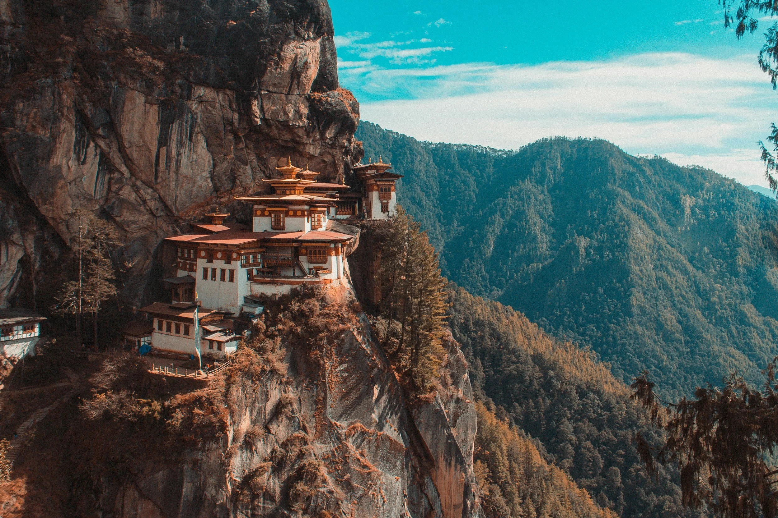 A monastery building constructed into a cliffside among forested mountains underneath a blue sky.