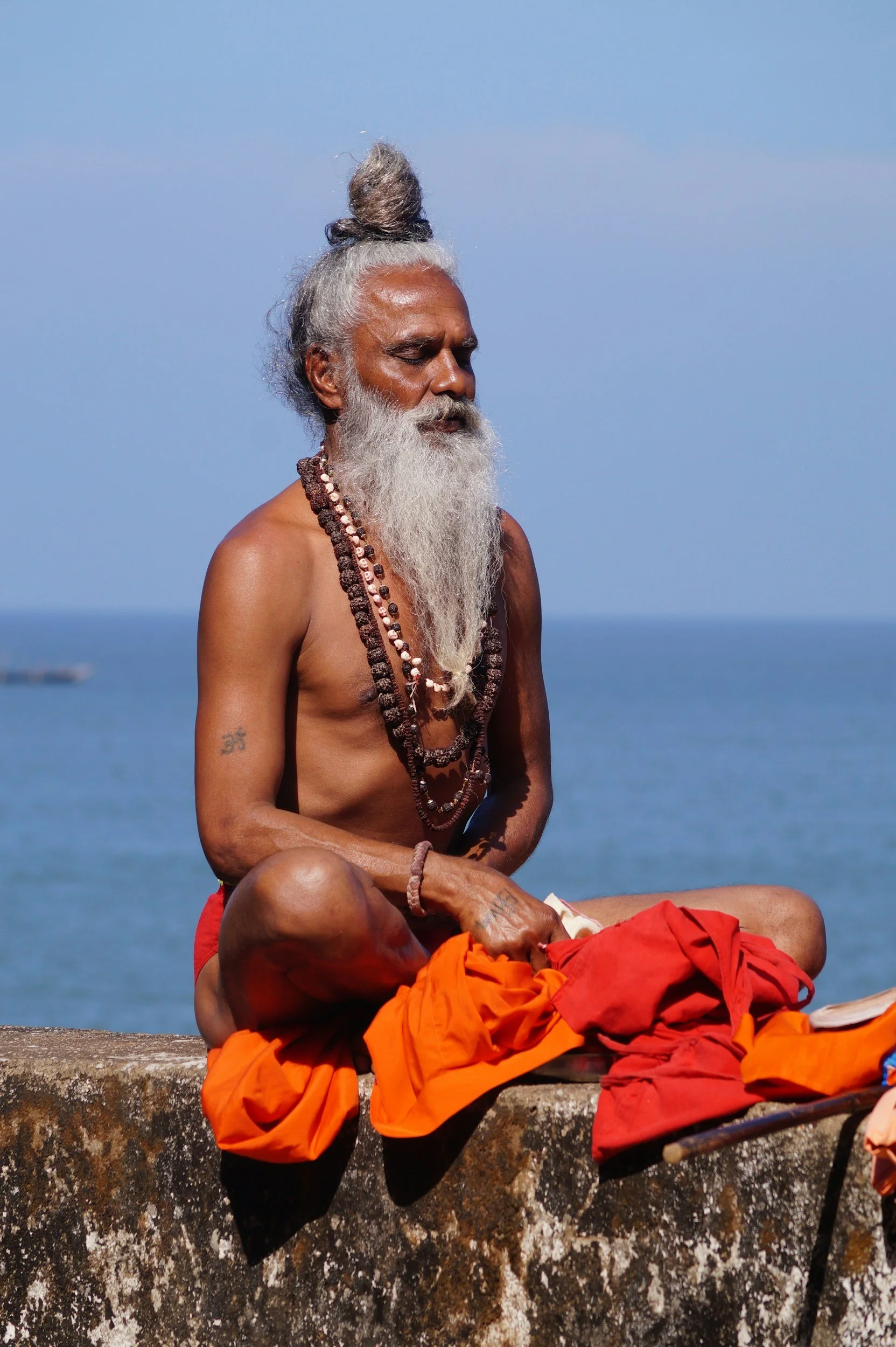 An elderly man with a long white beard, gray hair styled in a top knot, and dark skin, sits cross-legged in meditation with his eyes closed, wearing multiple beaded necklaces and red shorts, outdoors by the water. A monk dressed in orange robes rests with head on his lap.