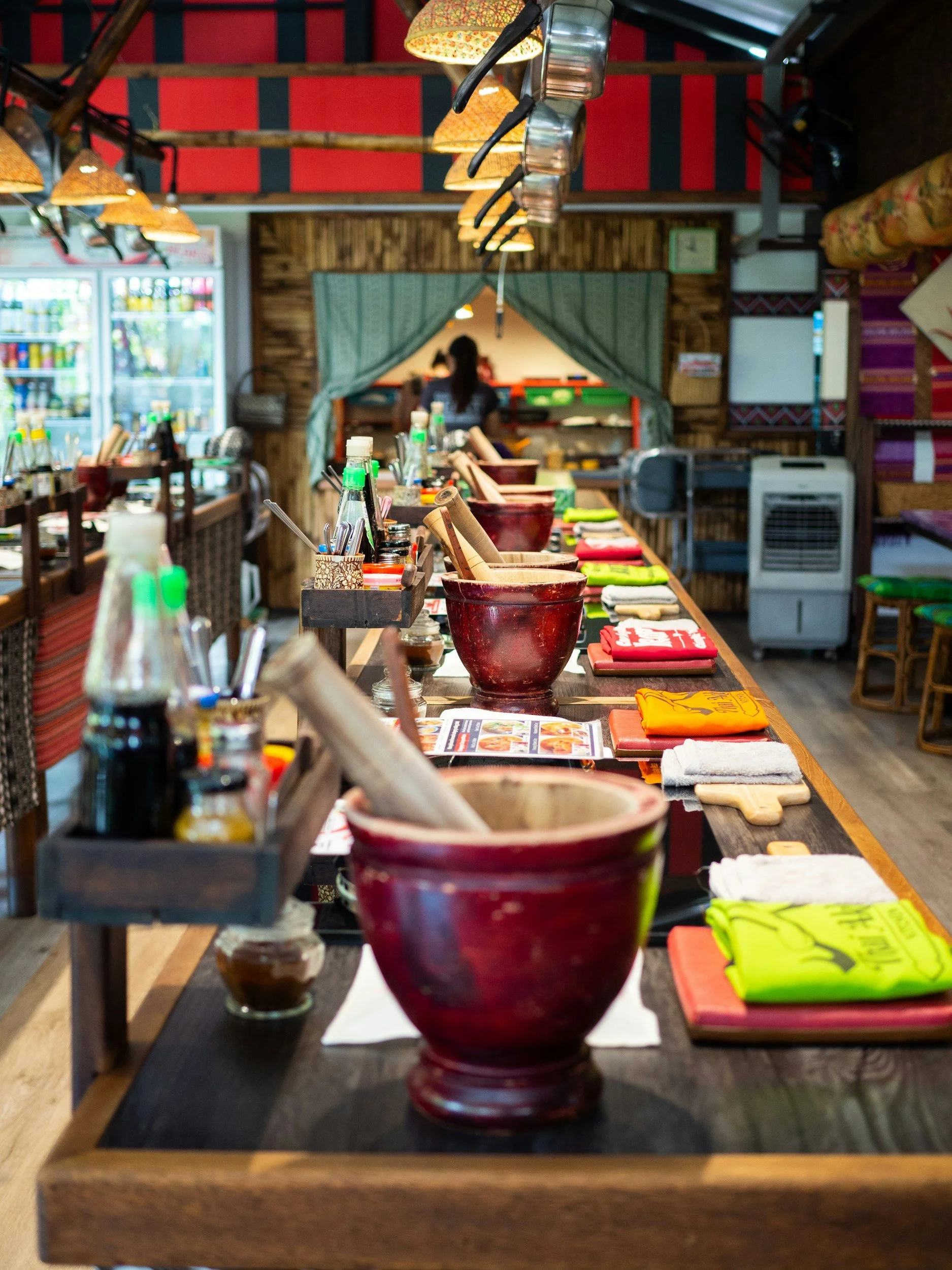 A row of red ceramic bowls on a black wooden table inside a restaurant, with various colorful napkins, utensils, and condiments in the background and a person sitting in the background.