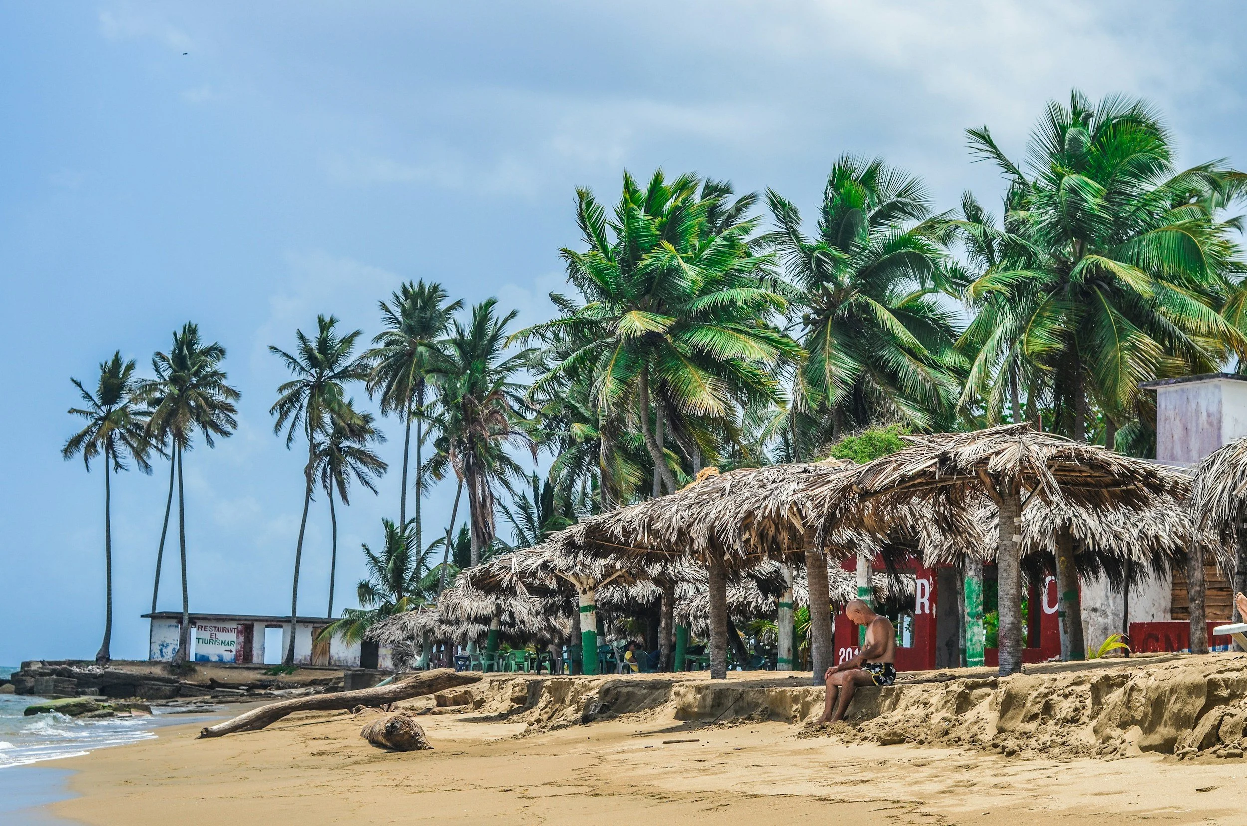 Beach scene with tall palm trees, thatched roof umbrellas, a person sitting on the sand, and a building in the background.