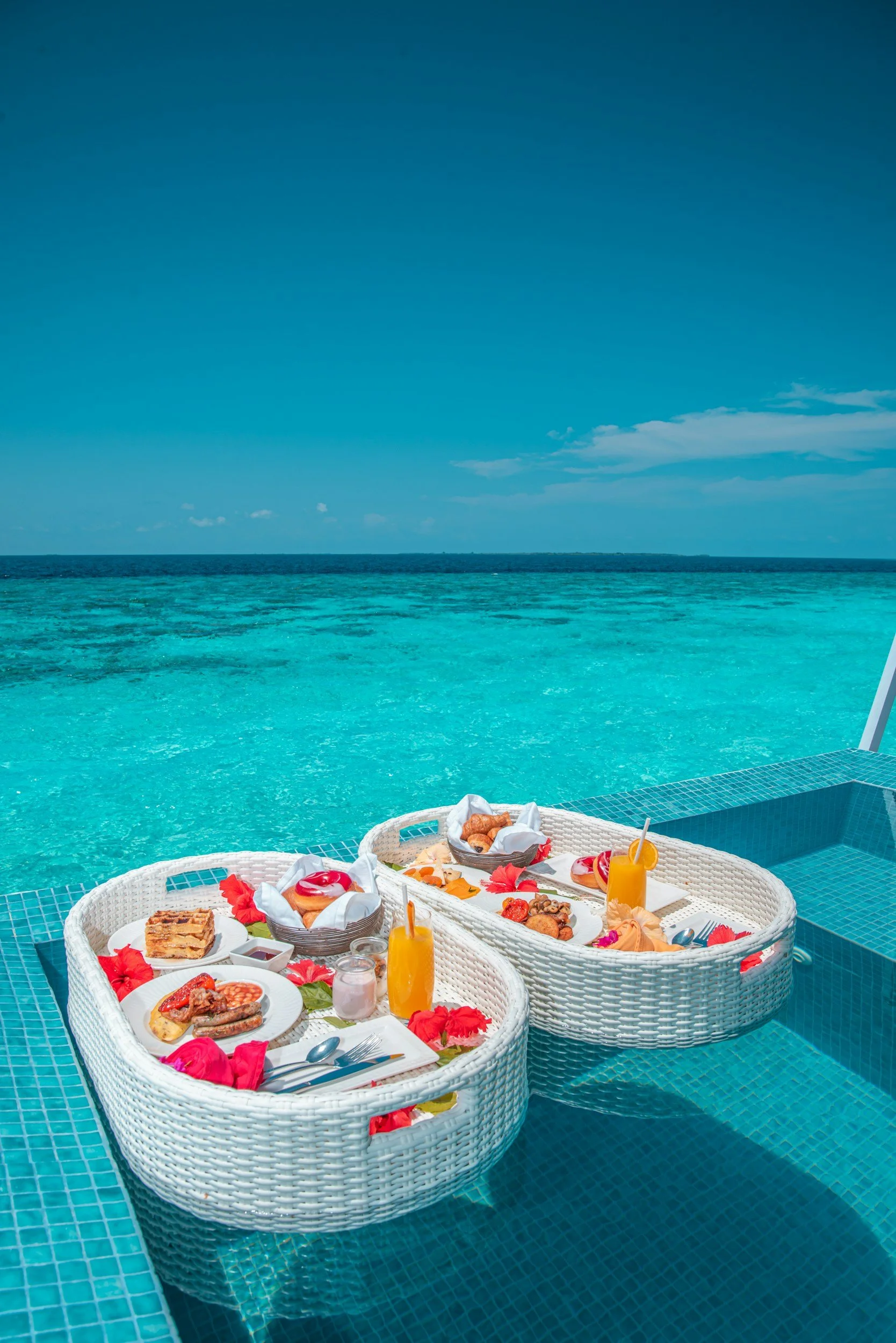 Breakfast setting with waffles, fruit, pastries, orange juice, and coffee on a poolside table overlooking the ocean and clear blue sky.
