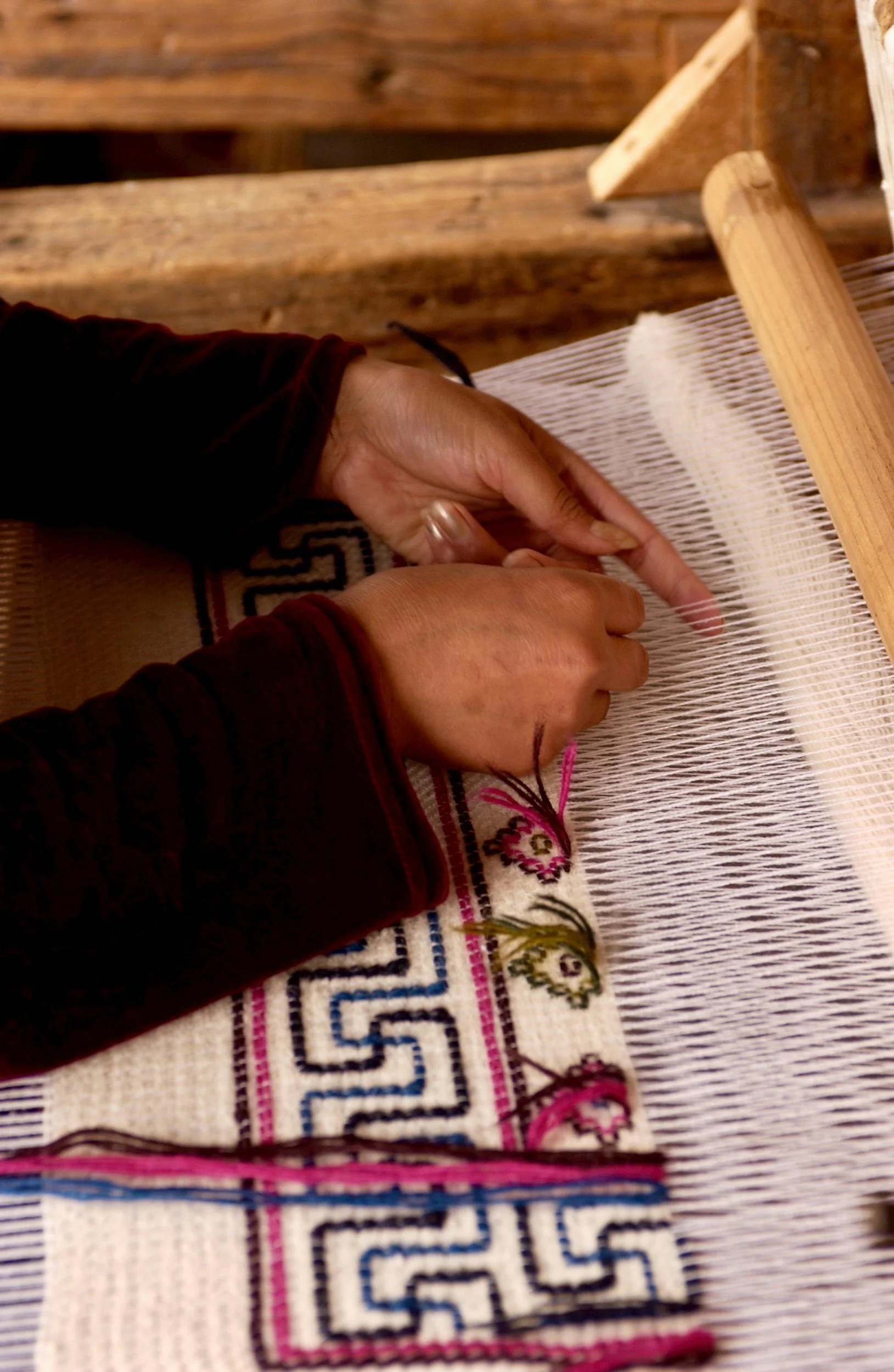 Person working on a woven textile on a wooden loom, with colorful embroidered fabric underneath.