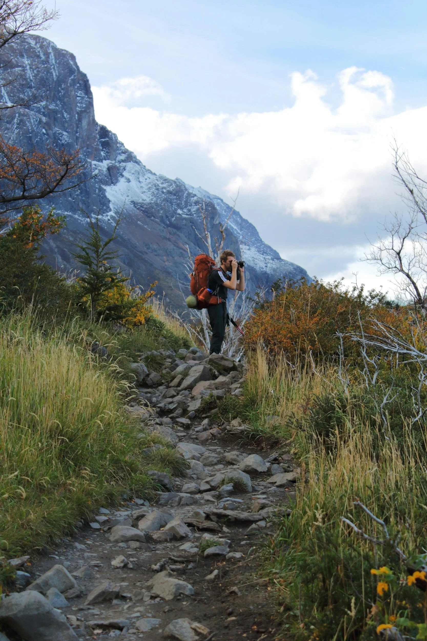 A hiker with a large orange backpack and camera taking a photo on a rocky trail in a mountainous landscape with snowy peaks, leafless trees, and a partly cloudy sky.