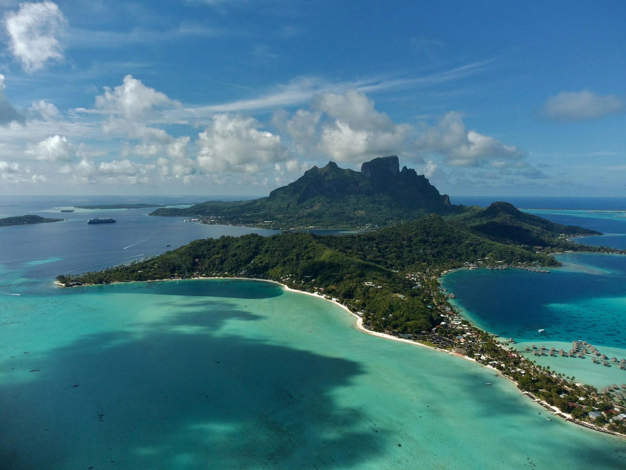 An aerial view of a tropical island with lush green mountains, overwater bungalows, and turquoise waters with boats and a cruise ship, under a partly cloudy sky.