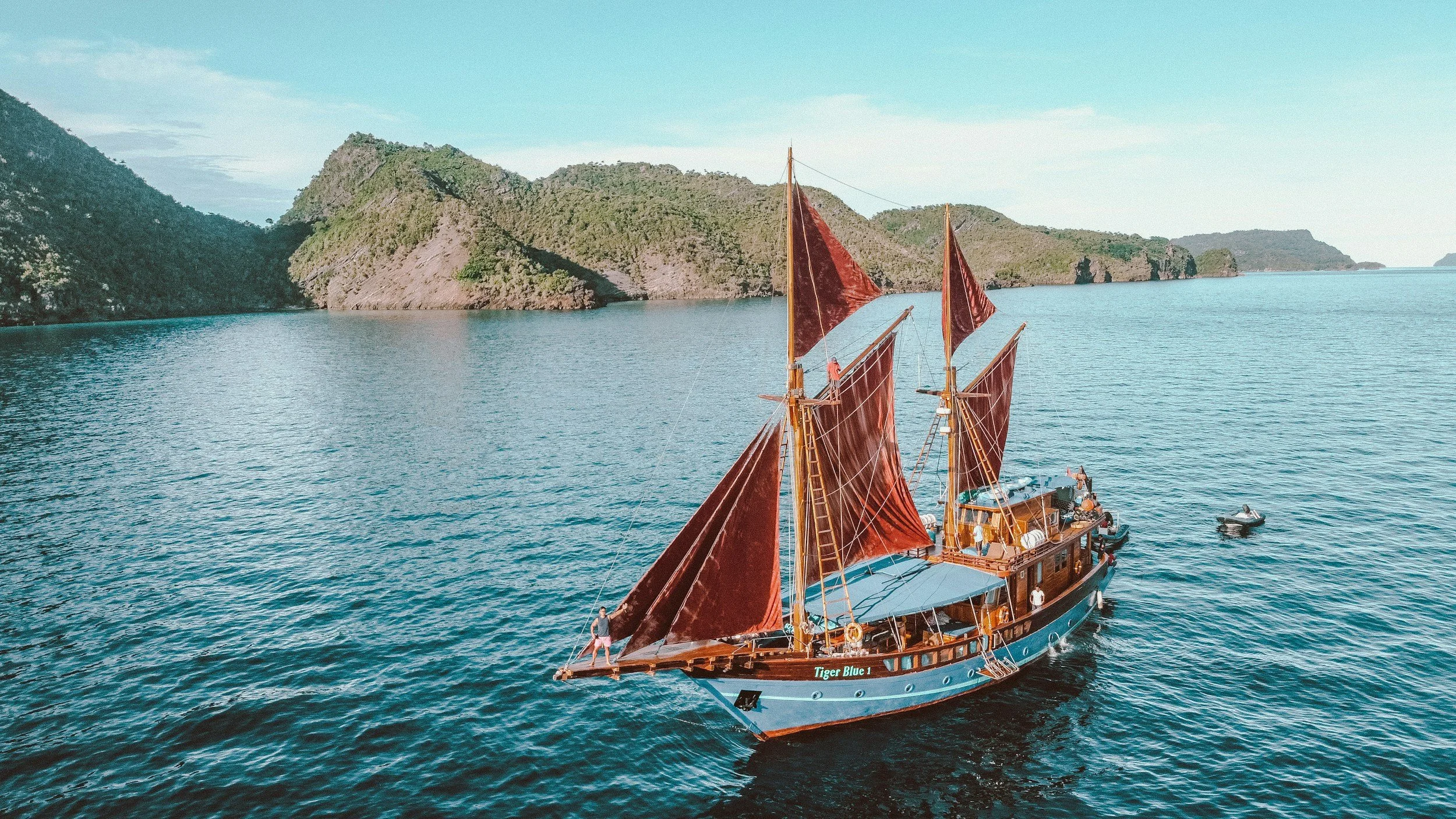 A detailed wooden sailing ship with red sails exploring a scenic ocean with lush green mountains and clear blue sky in the background.