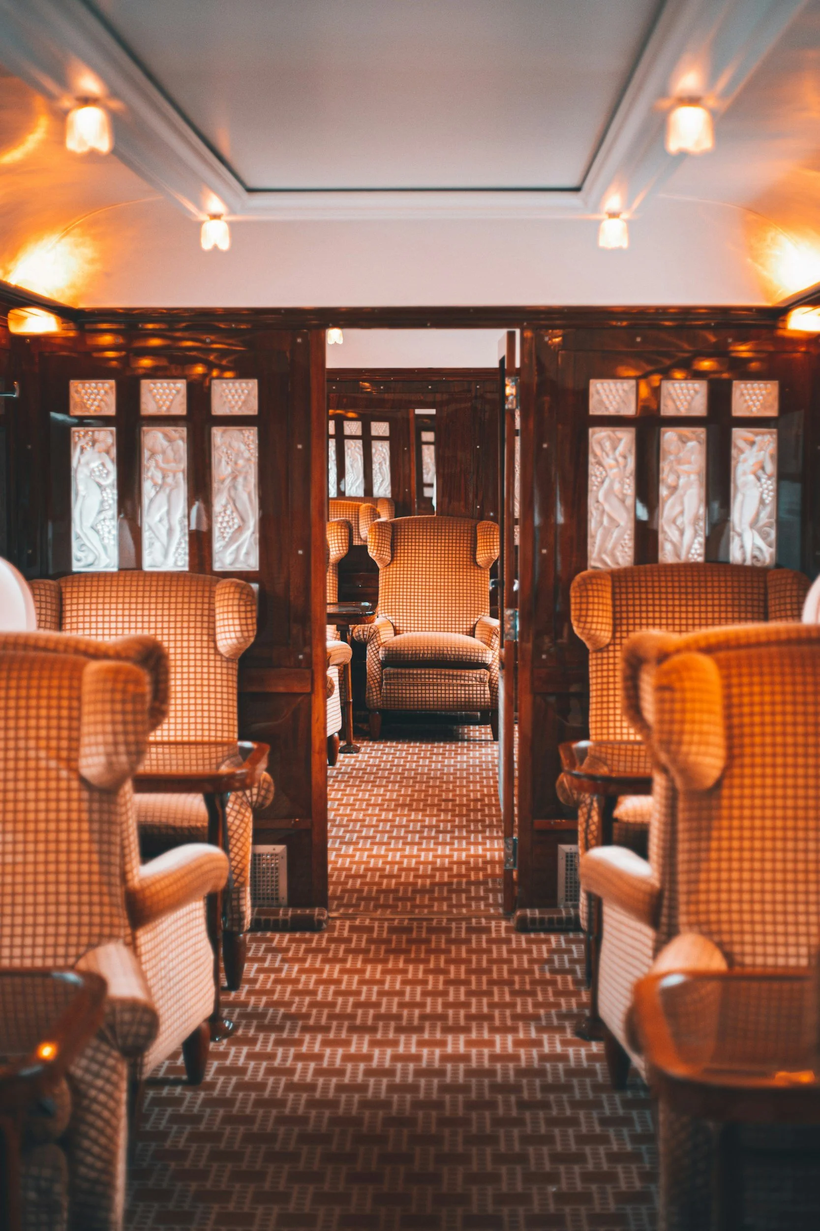 View of a cozy, vintage-style lounge with yellow checkered armchairs, dark wooden partition walls with carved reliefs, patterned carpet, and warm overhead lighting.