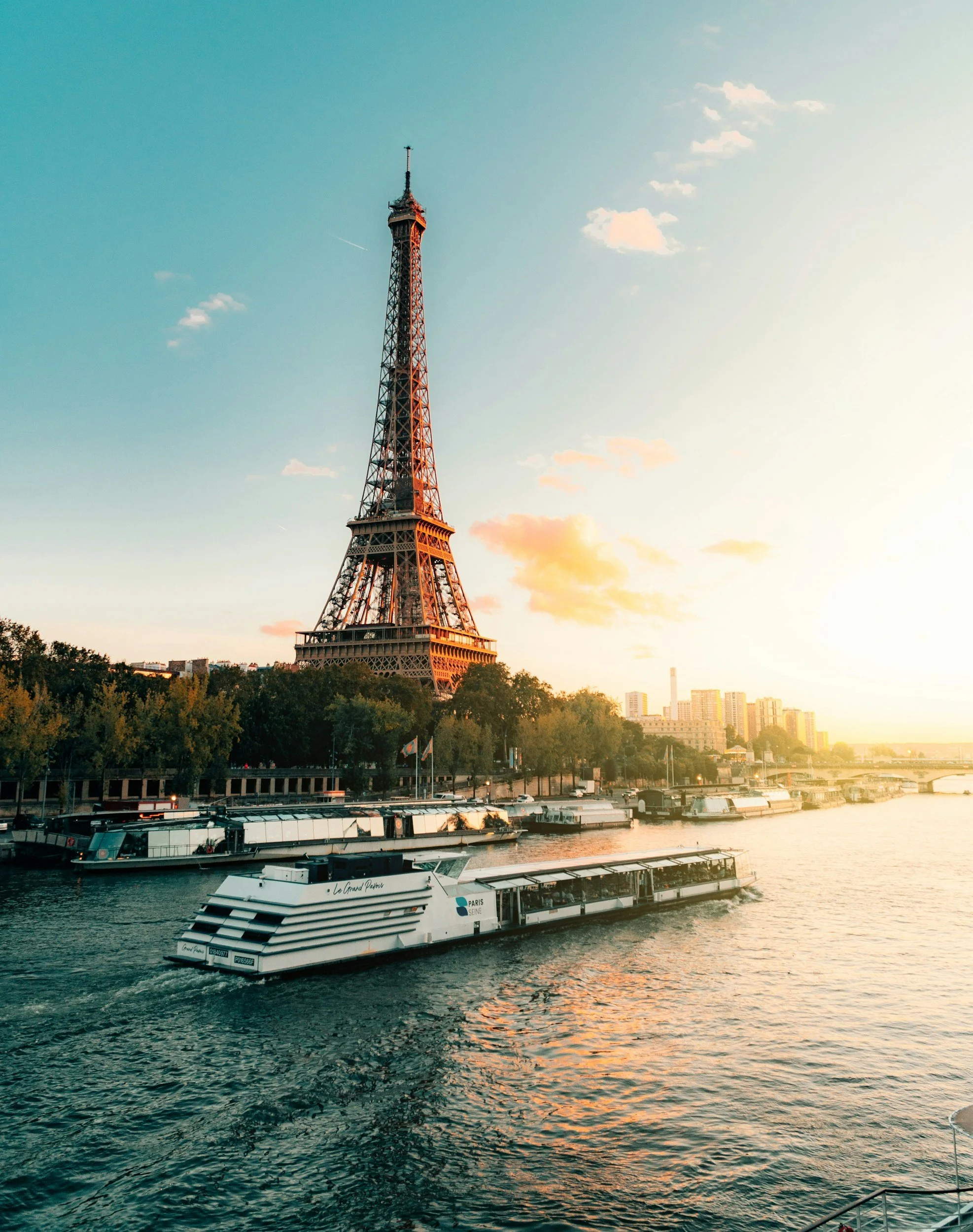 Paris Seine river cruise boat on the river Seine with the Eiffel Tower in the background at sunset, with buildings and trees along the riverbank.