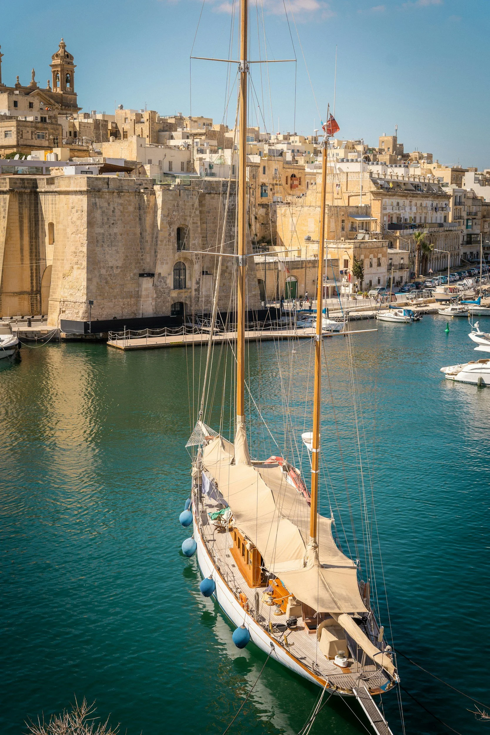 A sailboat docked in a harbor with historic stone buildings in the background, under a clear blue sky.