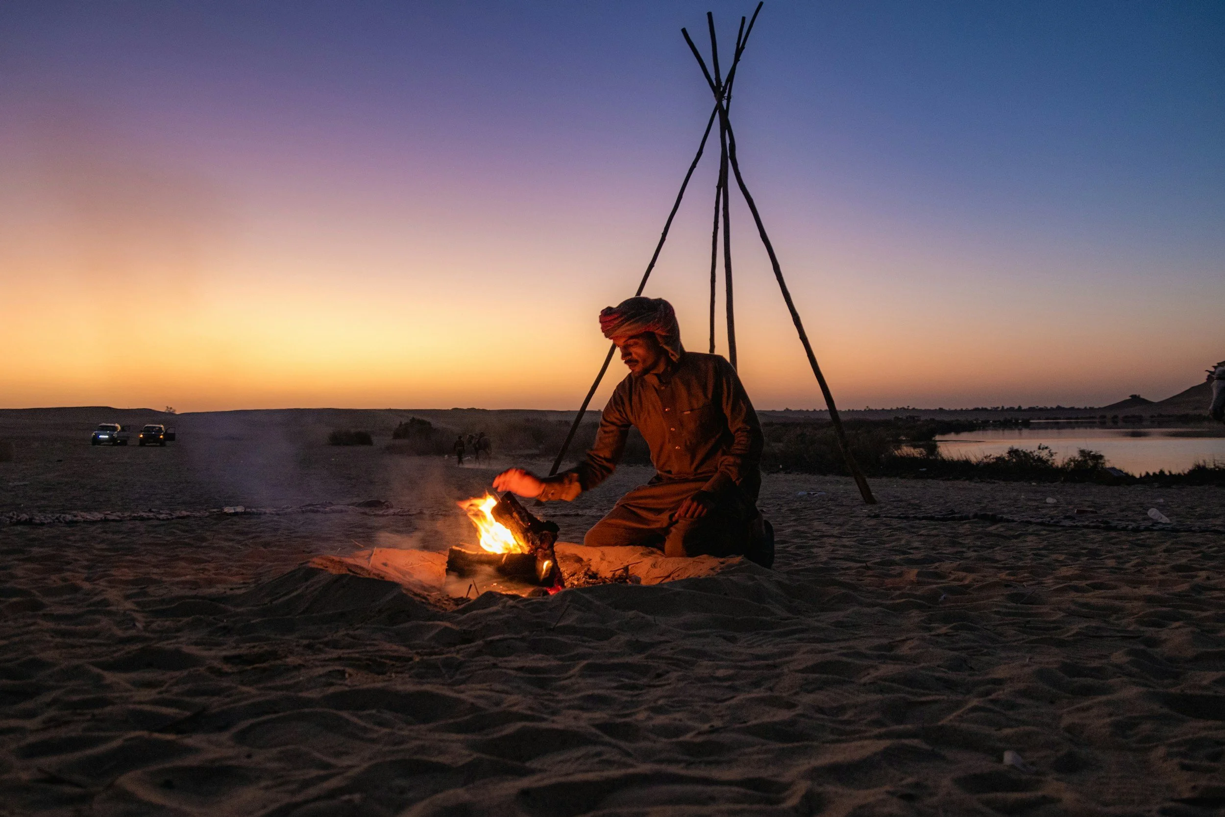 A man wearing traditional clothing preparing a fire on the desert ground during sunset, with a distant view of cars, a river, and hills.