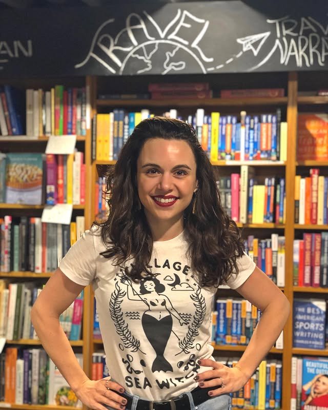 A woman with dark curly hair and red lipstick standing in front of a bookshelf at a bookstore, wearing a white t-shirt with a mermaid graphic and the words 'Village Sea Witch'.