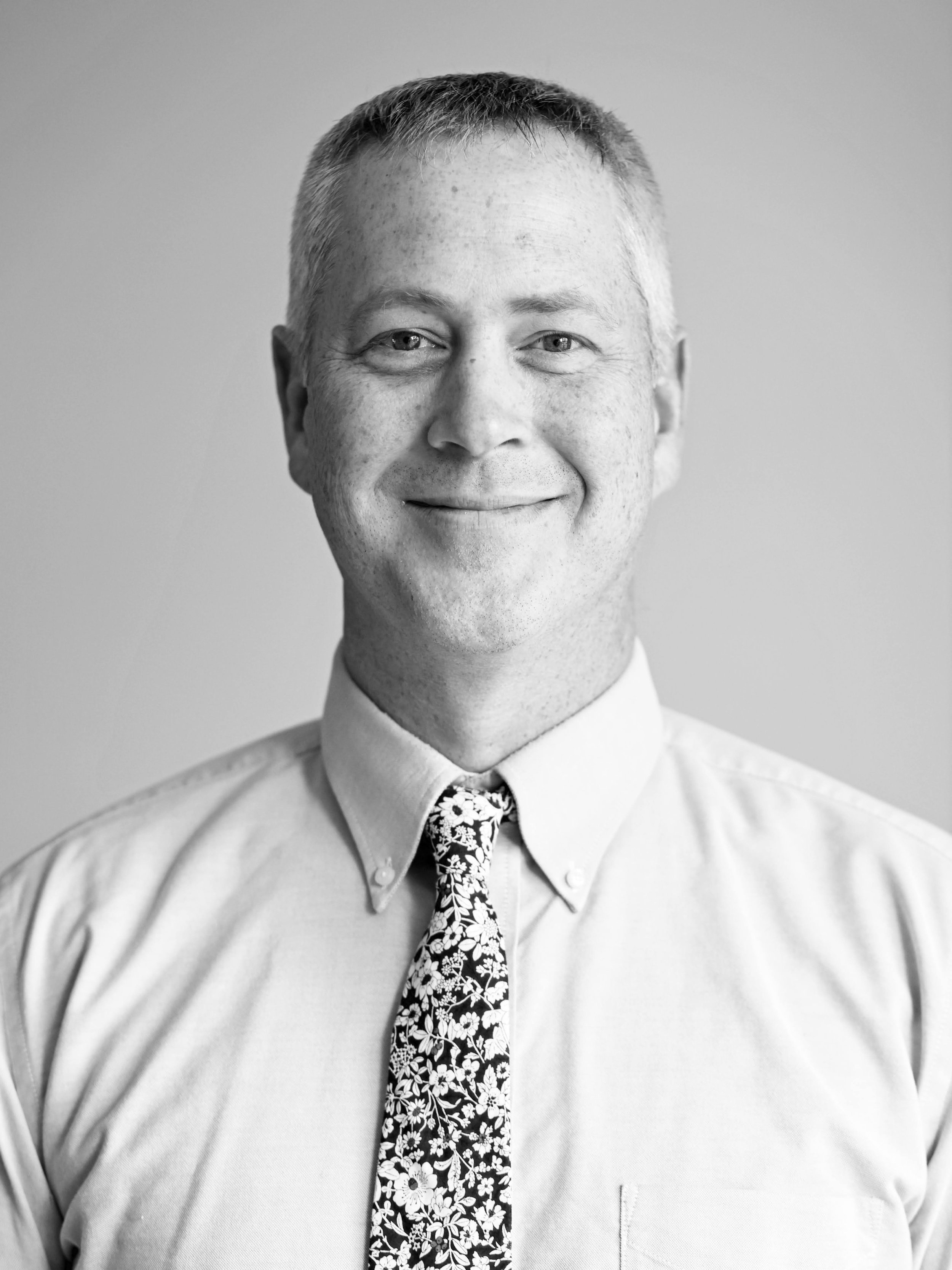 Black and white headshot of a smiling man with short, light-colored hair, wearing a collared shirt and a floral tie.