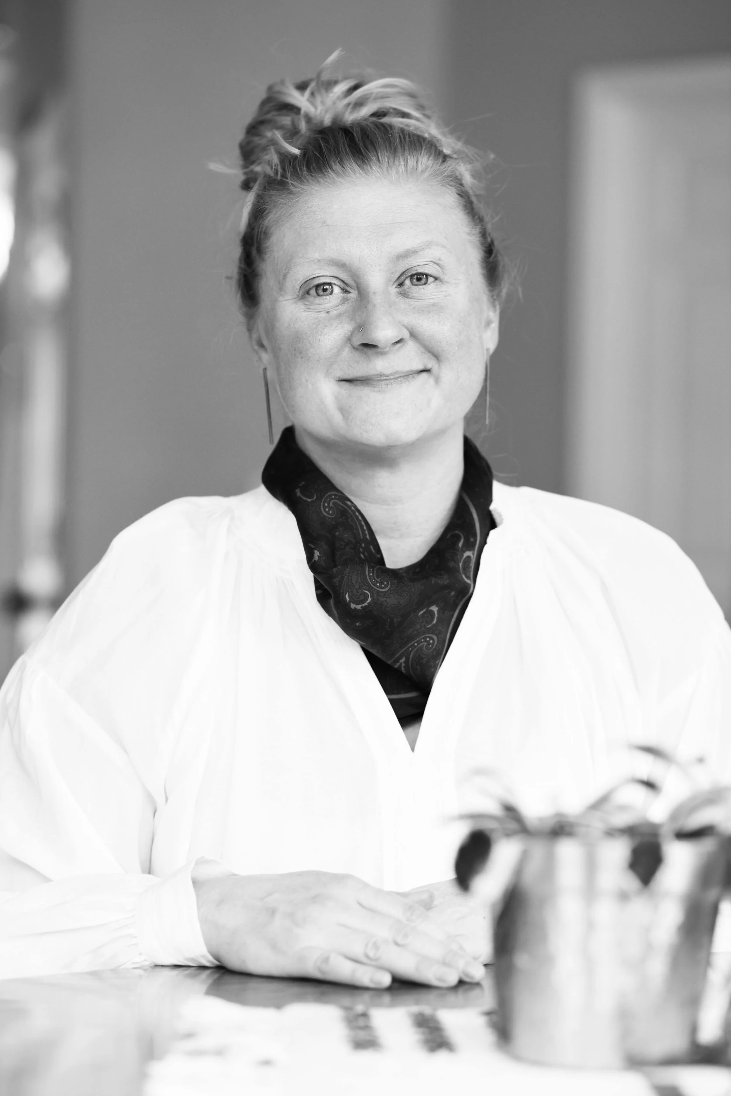 A woman with light skin, blonde hair tied up, wearing a white blouse, a dark patterned scarf, and hoop earrings, smiling while sitting at a table with a plant.